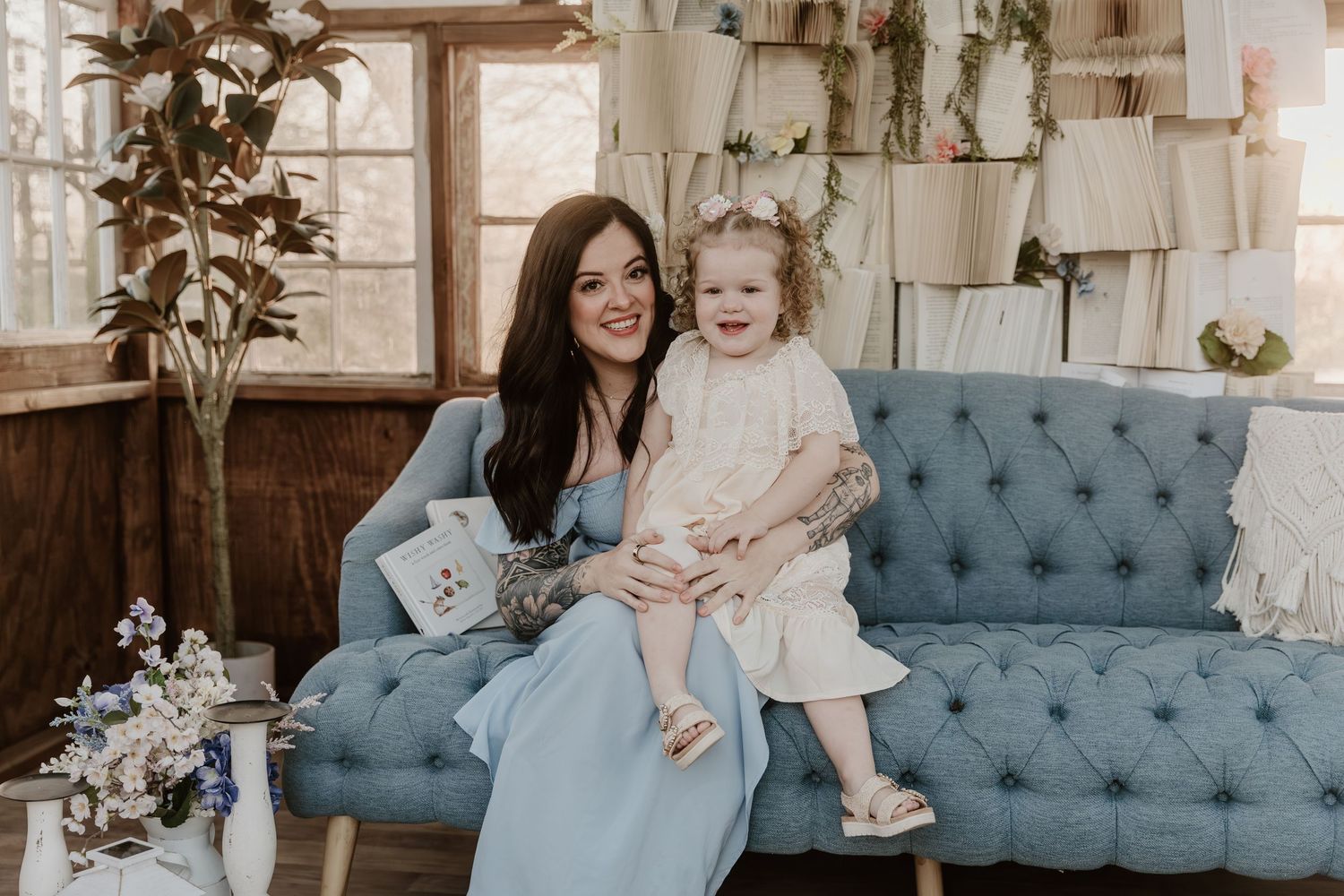 Woman in blue dress sitting on teal sofa holding smiling toddler girl in white dress in decorated room.