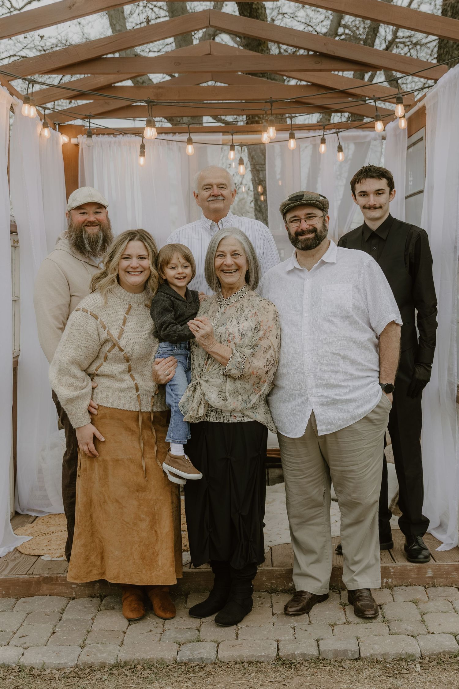 Family group photo in neutral earth-toned outfits standing against white wall under rustic wooden pergola.