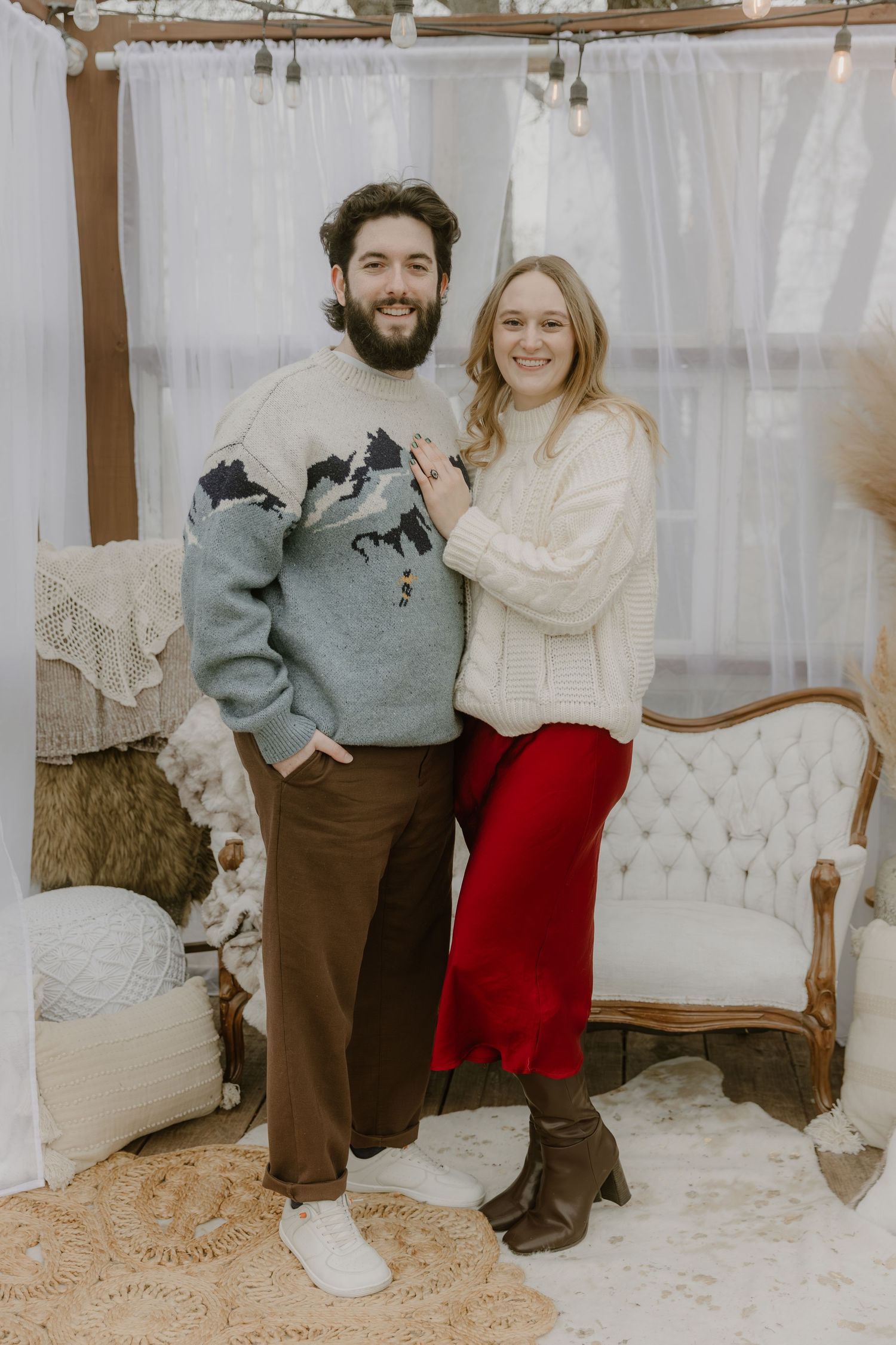 A couple in casual attire poses together in front of a white tufted sofa in a cozy indoor setting.