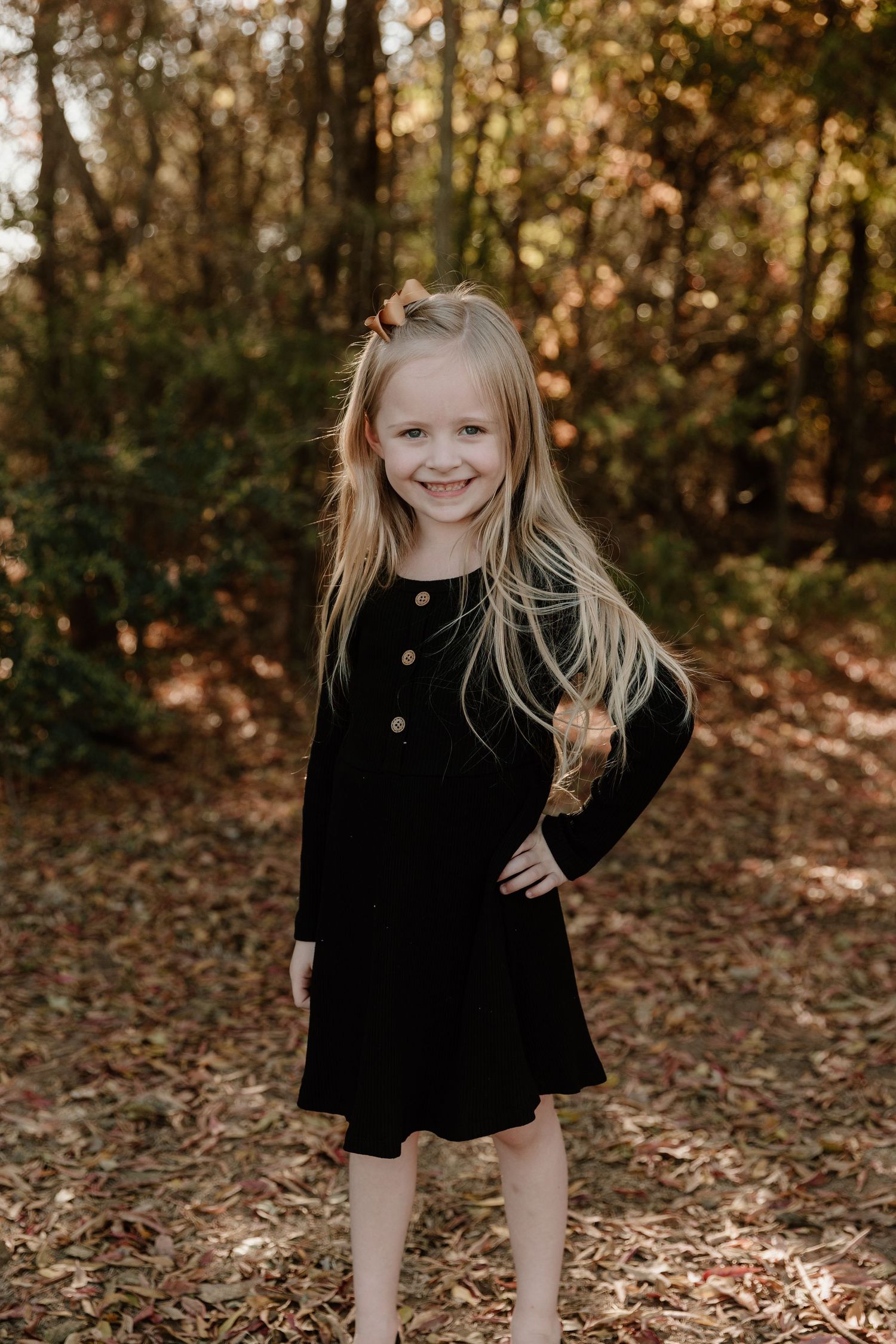 A child in a black dress poses confidently on fallen autumn leaves in a wooded setting during golden hour.