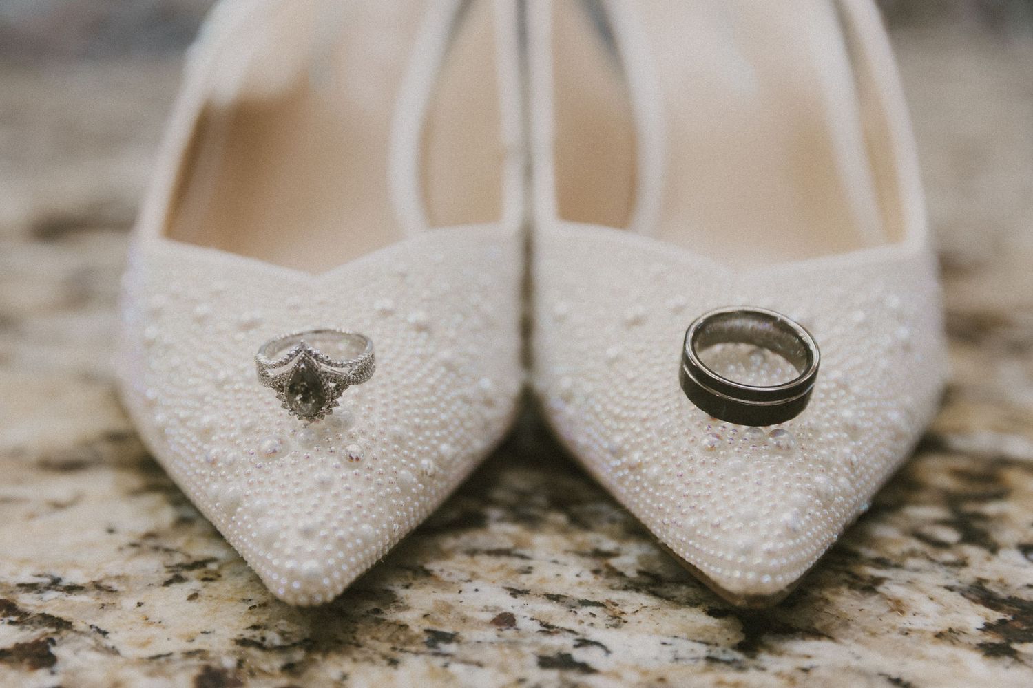 White beaded bridal heels with diamond engagement ring and gold wedding band displayed on marble surface.