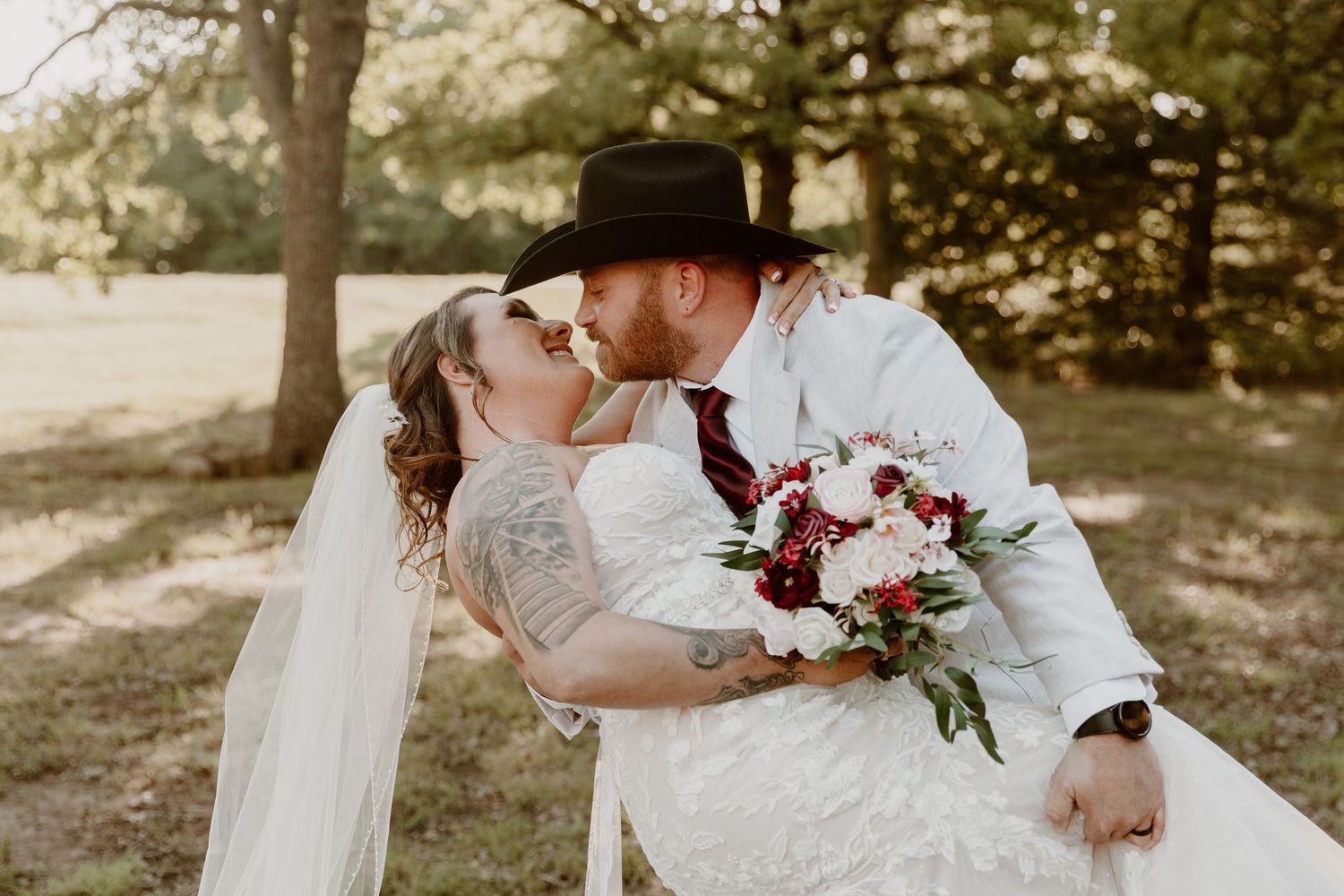 A romantic wedding couple sharing a kiss in a nature setting while holding a burgundy and white floral bouquet.