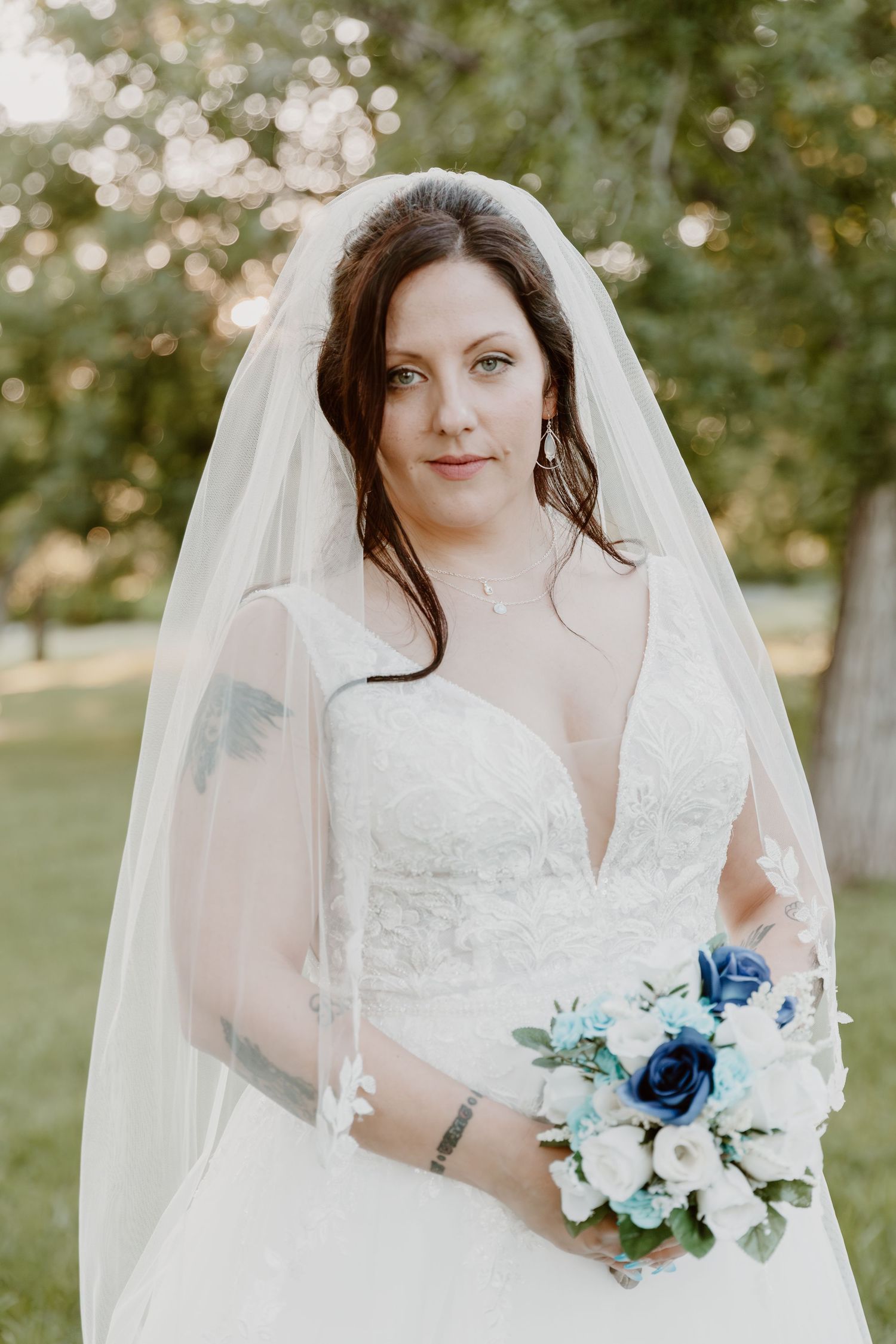 A bride in a white lace wedding dress holds a blue and white bouquet during an outdoor summer ceremony.