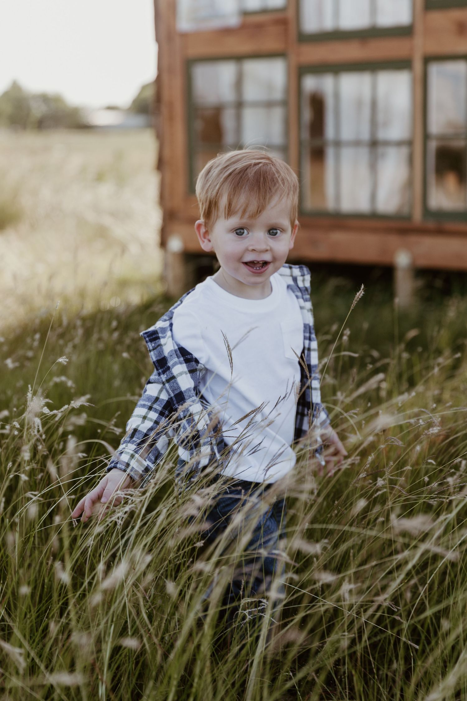 A child sits among tall grass in a rustic setting with brown buildings visible in the background.