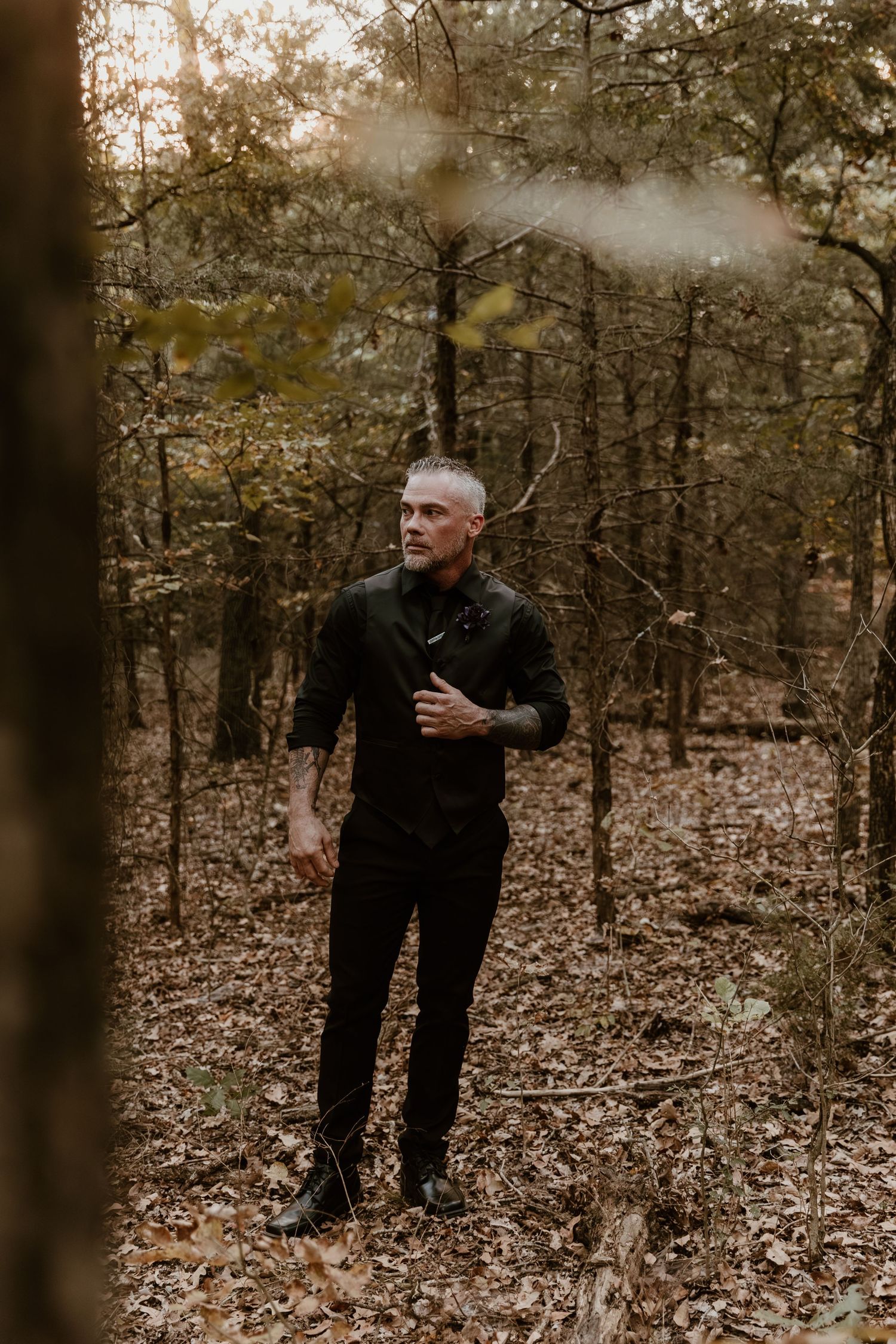 A person in dark clothing sits contemplatively in an autumn forest setting with fallen leaves on the ground.