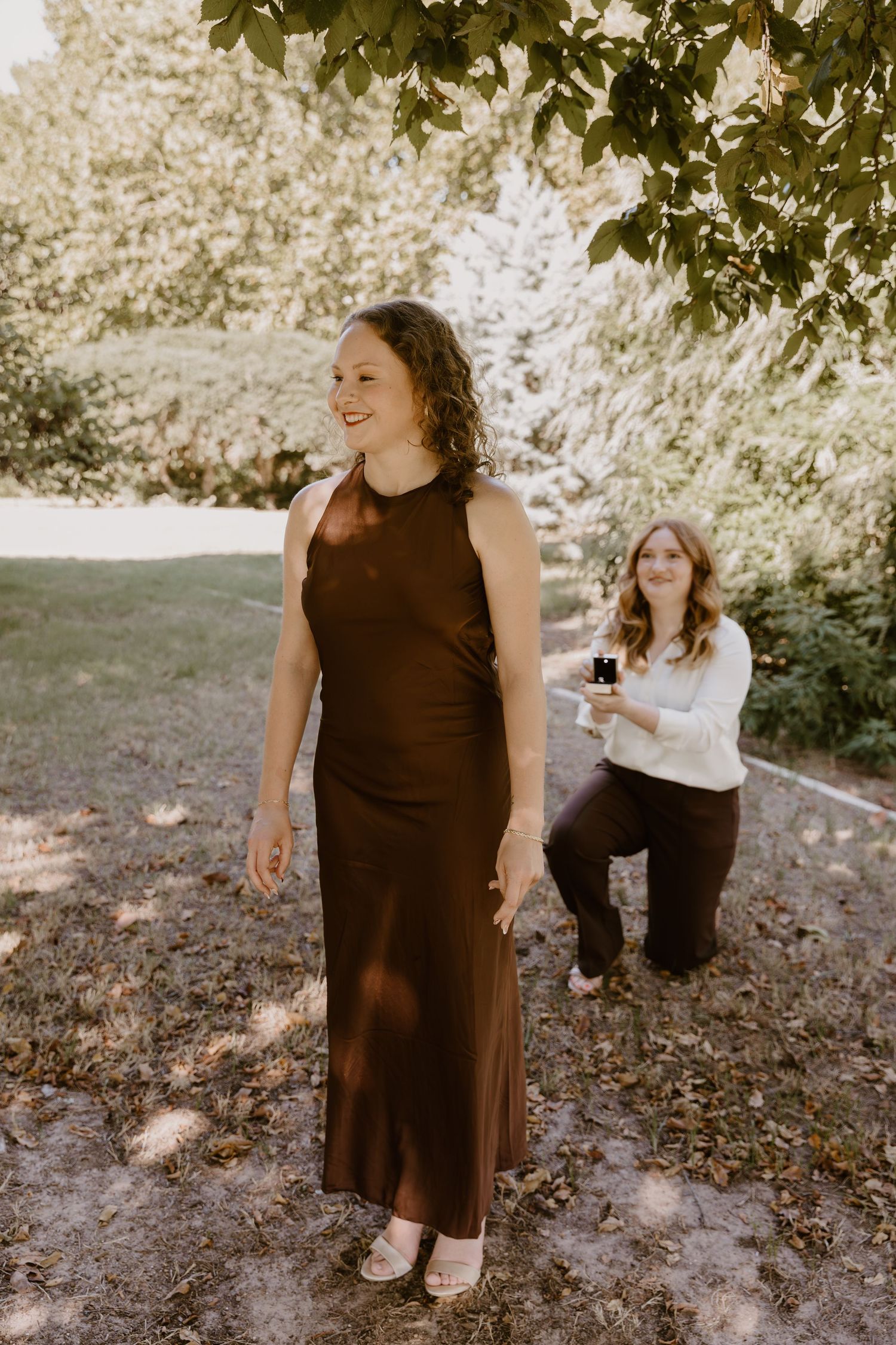 Two people share a joyful moment outdoors, one wearing a brown formal dress while another kneels in white attire.