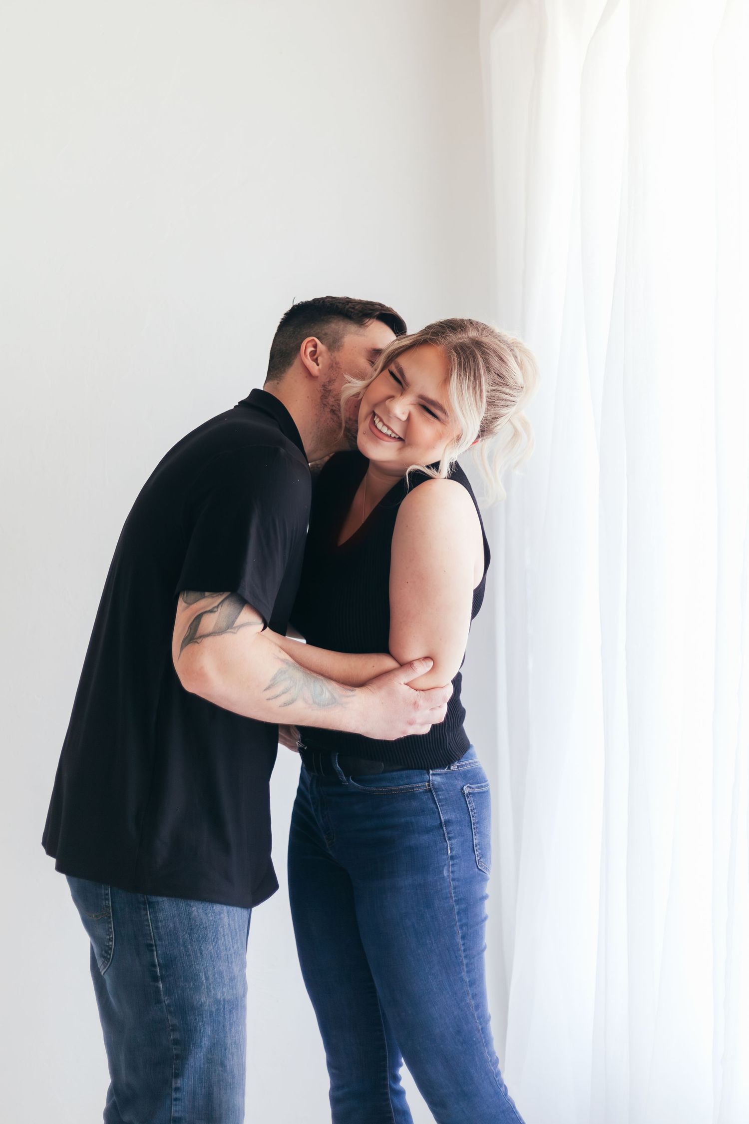 Intimate moment captured between couple against white window backdrop.