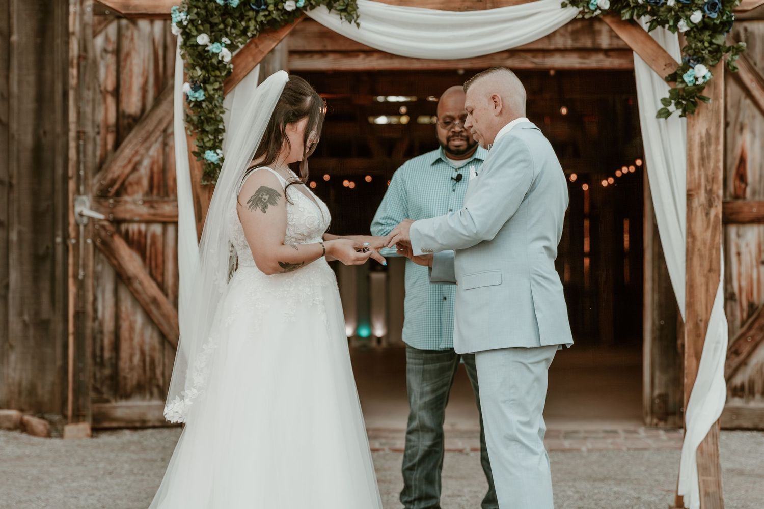 A couple exchanges rings during a rustic barn wedding ceremony decorated with white drapes and string lights.