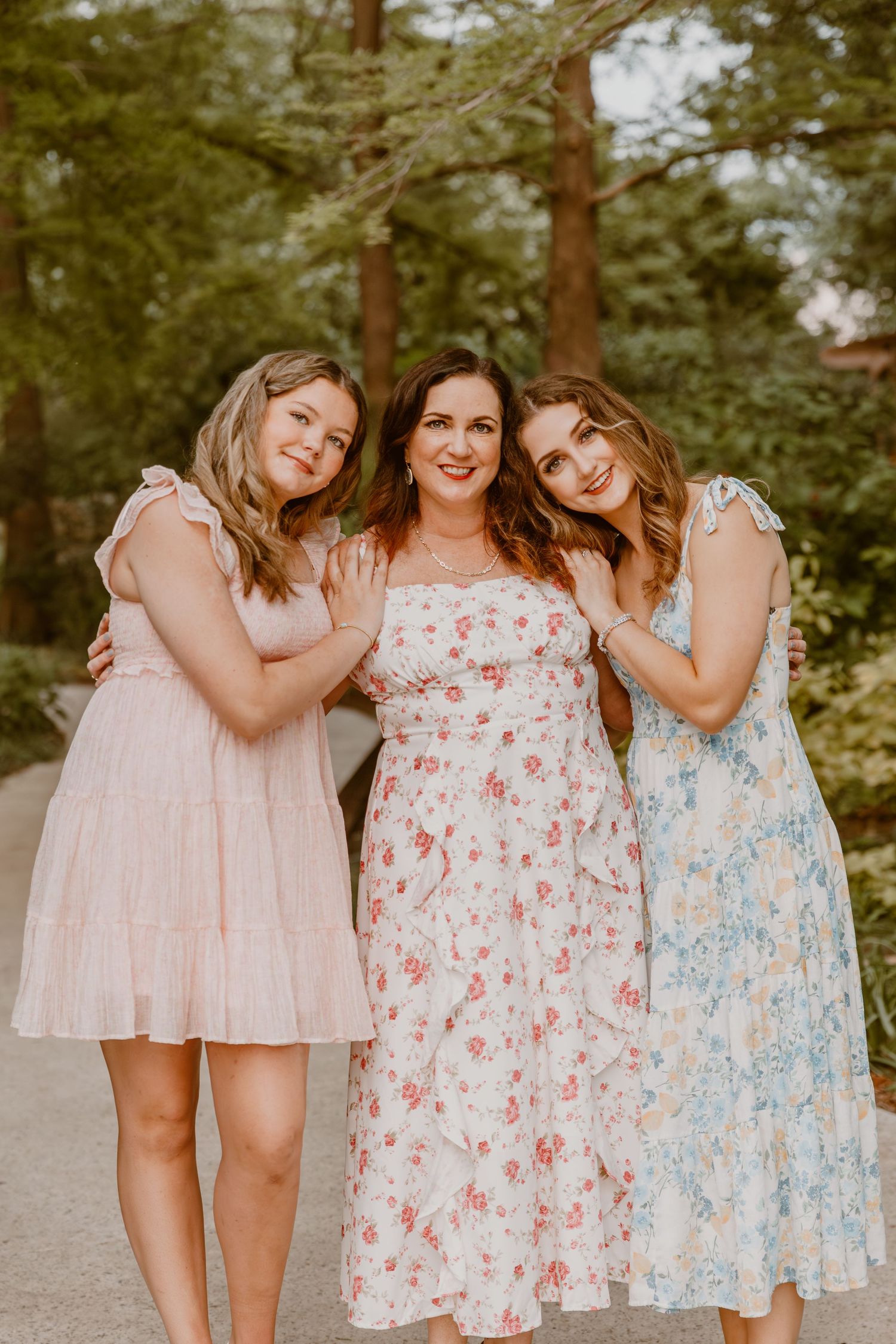 Three friends pose together outdoors wearing romantic floral summer dresses in pink, red and blue tones.