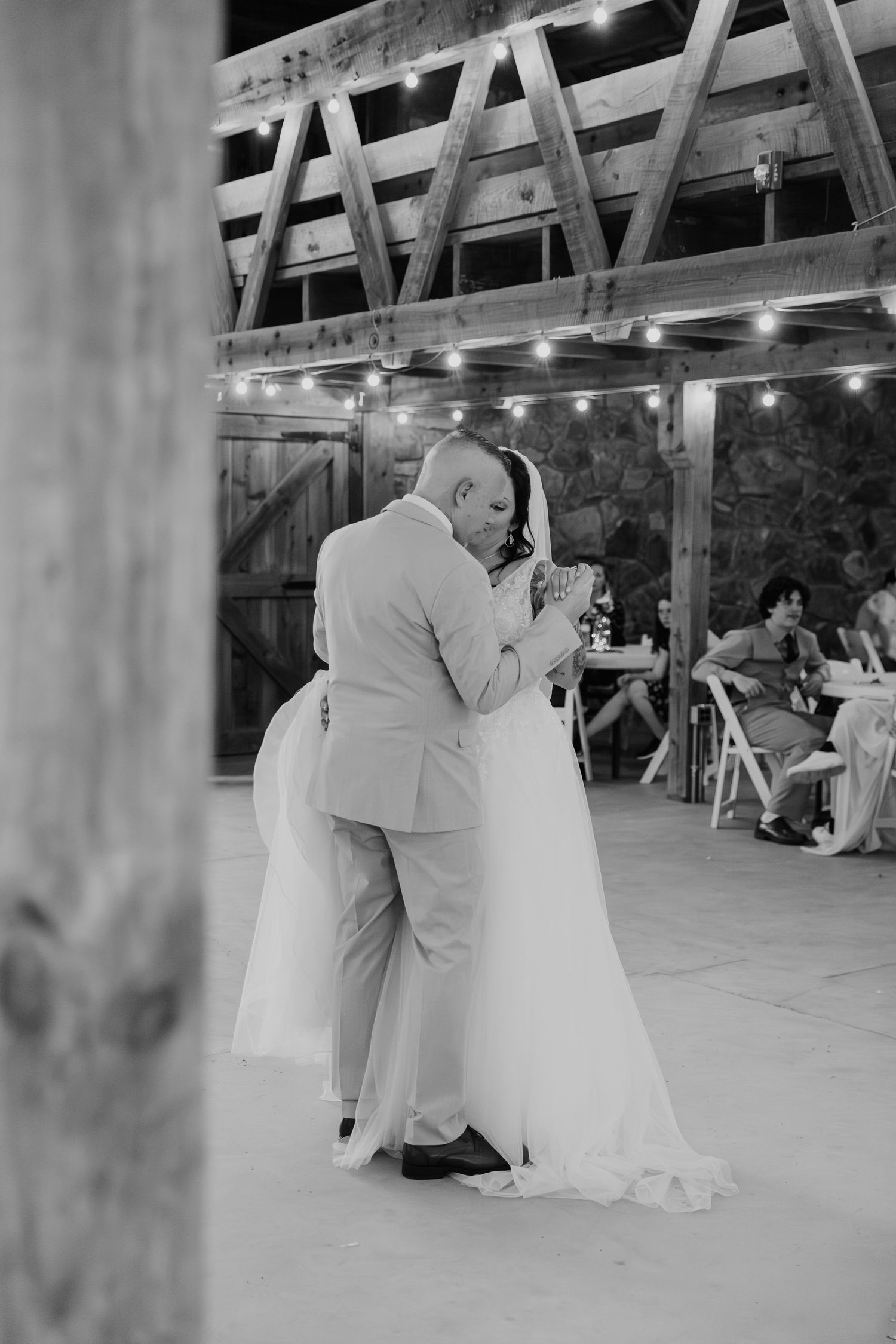 A newly married couple shares their first dance together in a rustic barn venue with string lights overhead.