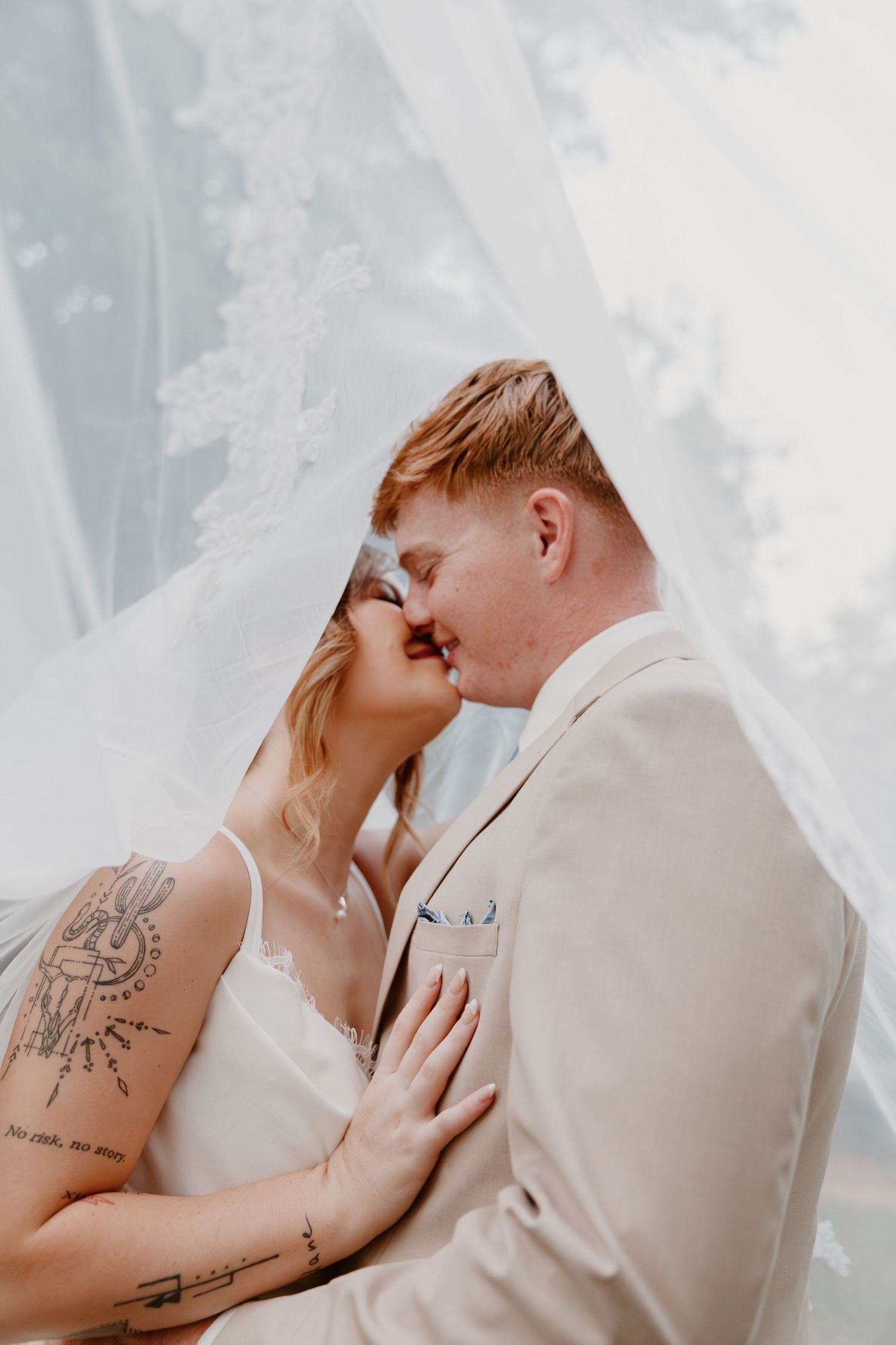 Romantic wedding portrait shows couple sharing an intimate moment under flowing white veil outdoors.