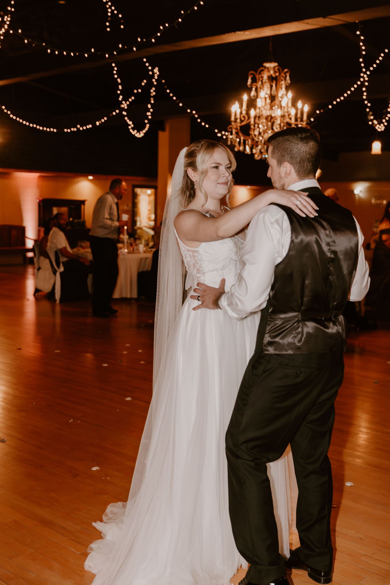 A couple shares their first dance at a wedding reception under twinkling string lights and an elegant chandelier.