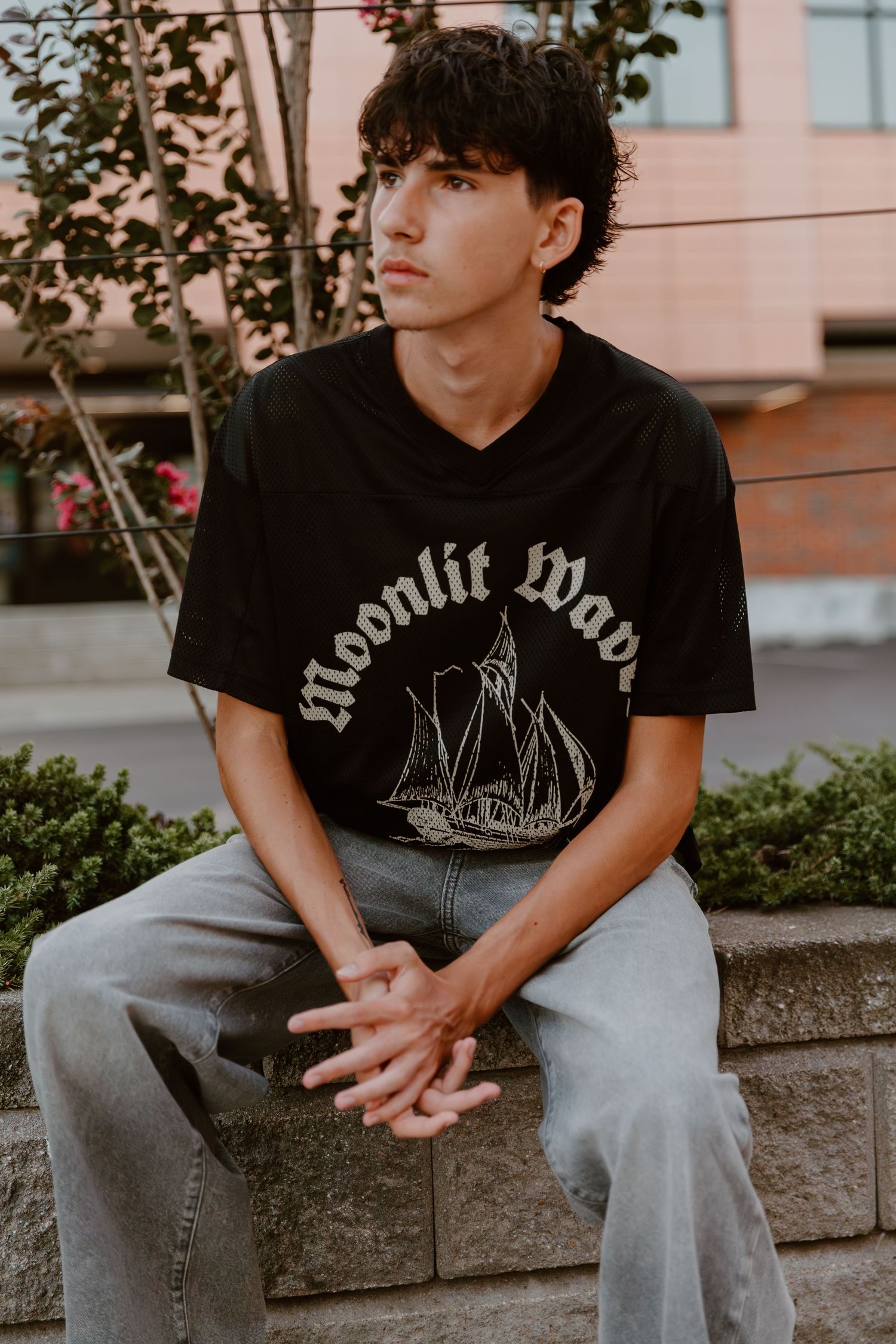 Person wearing black band t-shirt and jeans sits on stone ledge in urban setting at sunset.