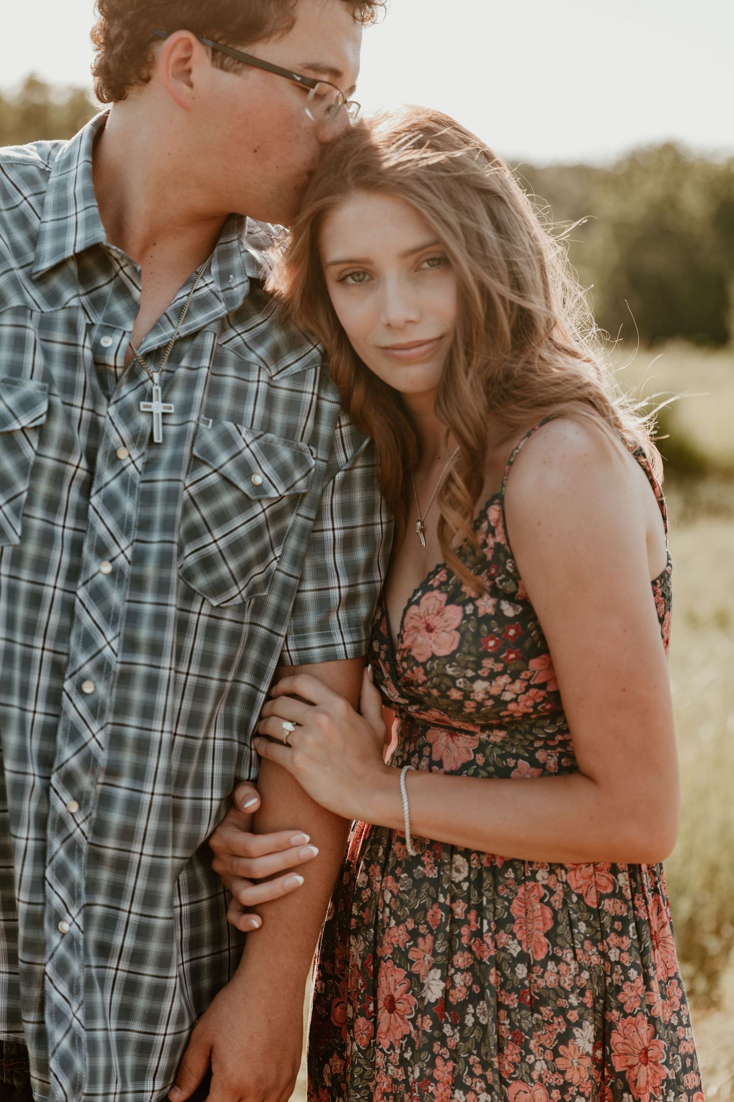 A couple embraces in warm sunlight, wearing a plaid shirt and floral dress while standing in a natural outdoor setting.