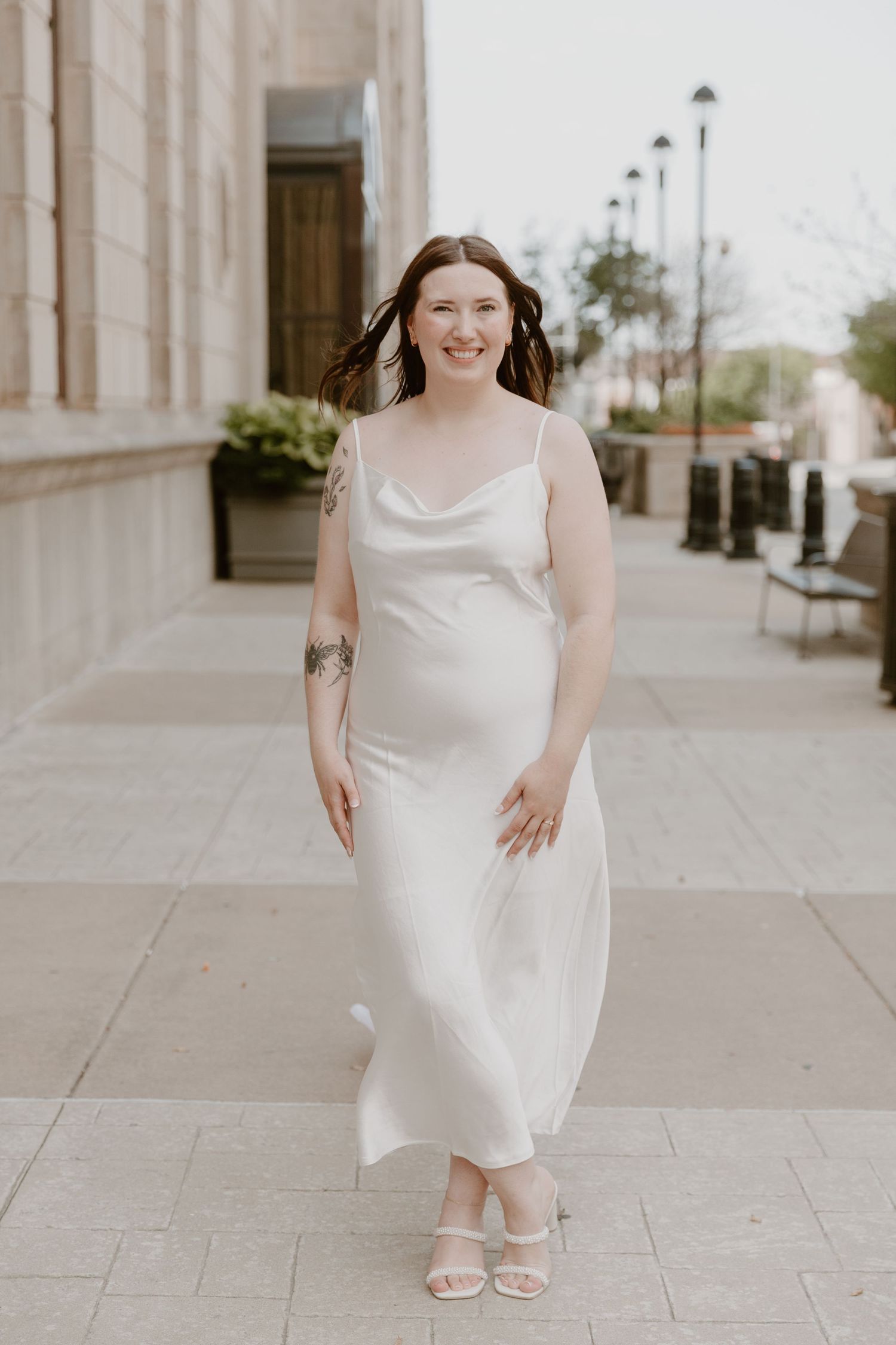 A modern bridal look featuring a minimalist white dress and sandals against a classic stone column backdrop.