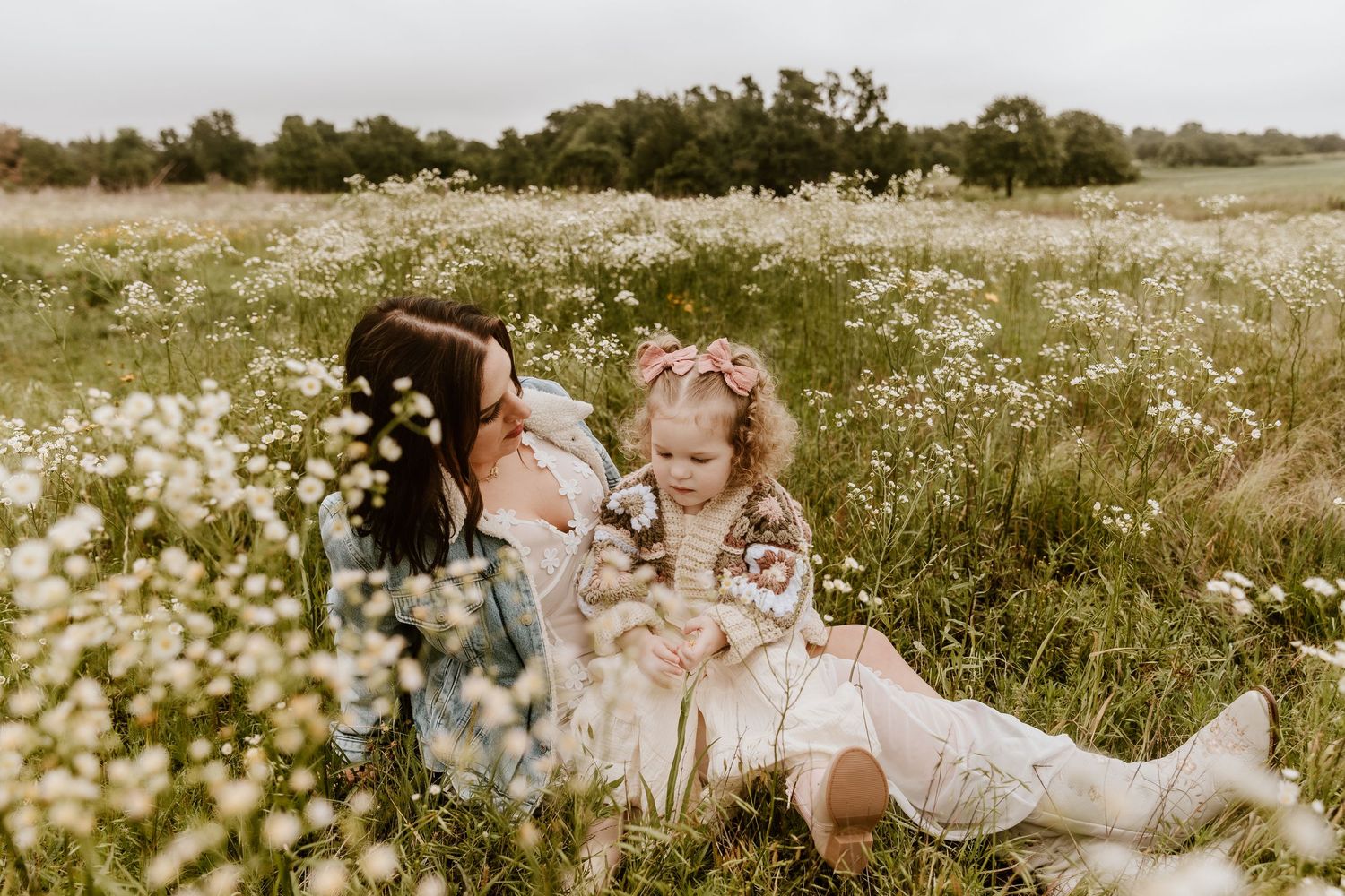 Mother and toddler daughter sitting together in a wildflower field, sharing a tender moment among white blooms.