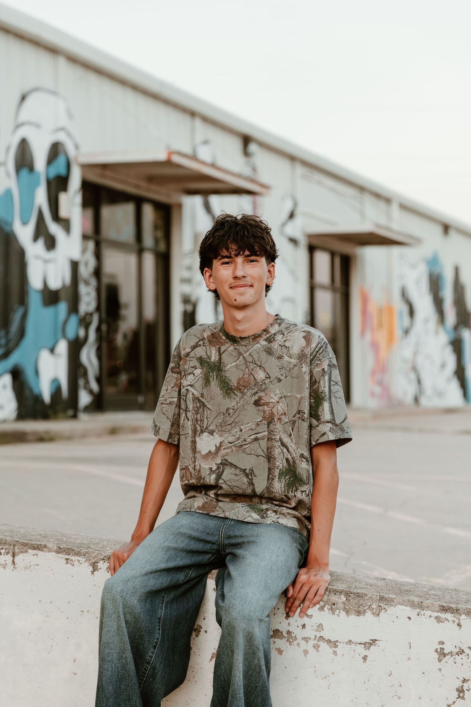 A person in jeans and a grey t-shirt sits on a concrete step in front of a building with painted murals.