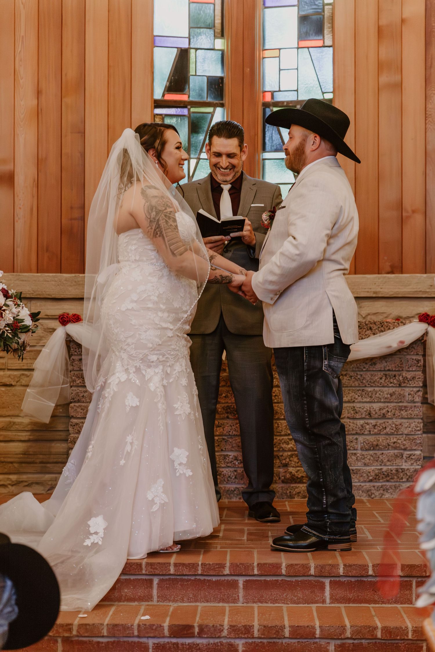 Wedding ceremony takes place in front of a stained glass window on brick steps with wooden paneled walls.