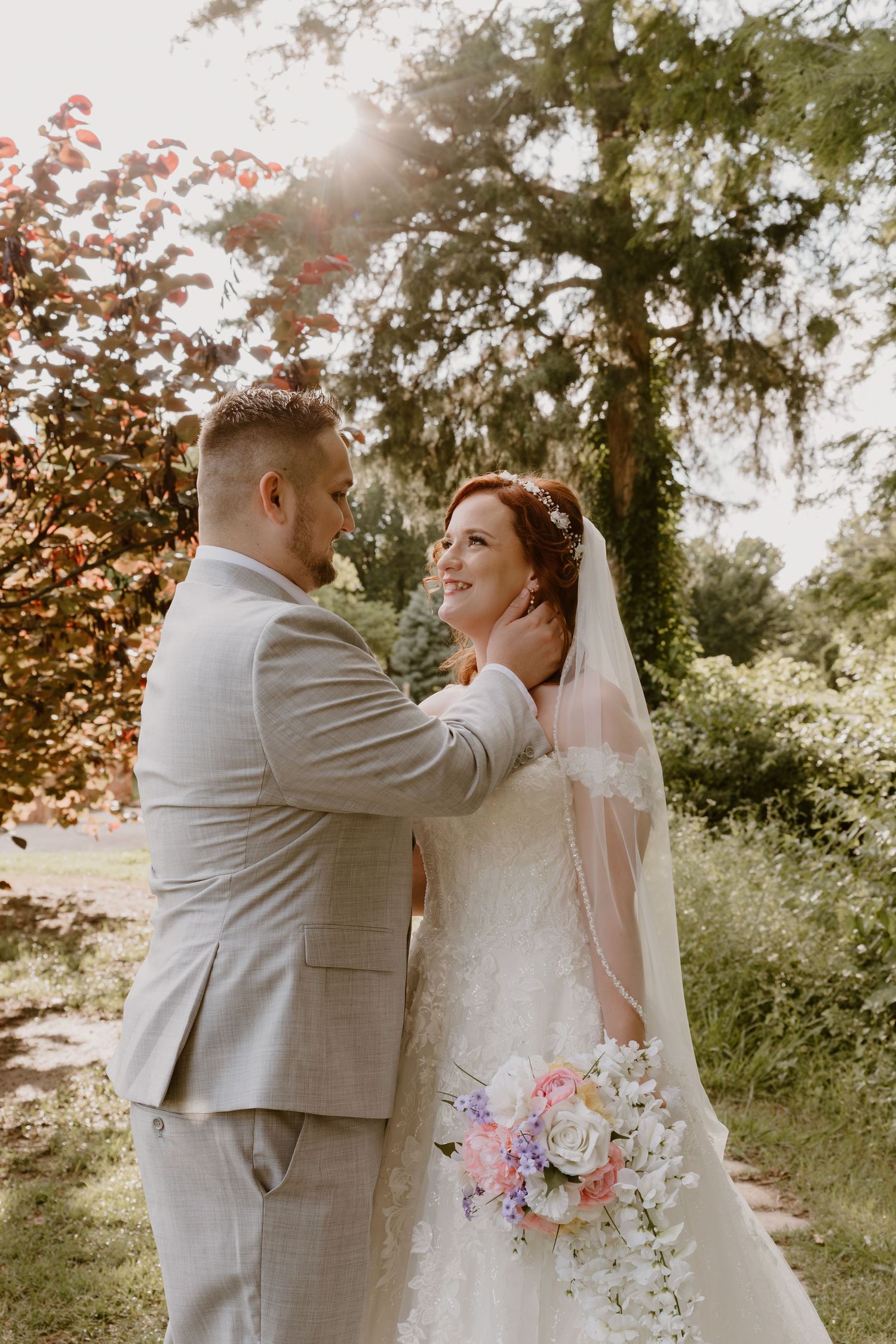 A tender wedding moment captured outdoors with sunlit trees, flowing veil and pastel floral bouquet in a natural setting.