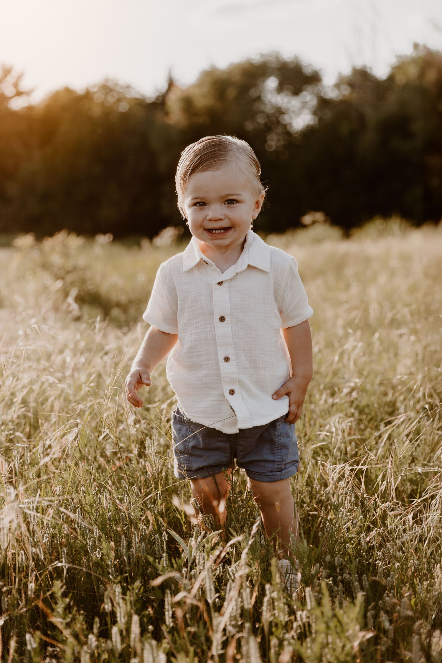 Child in white button-down shirt and denim shorts stands in golden sunlit field during sunset.