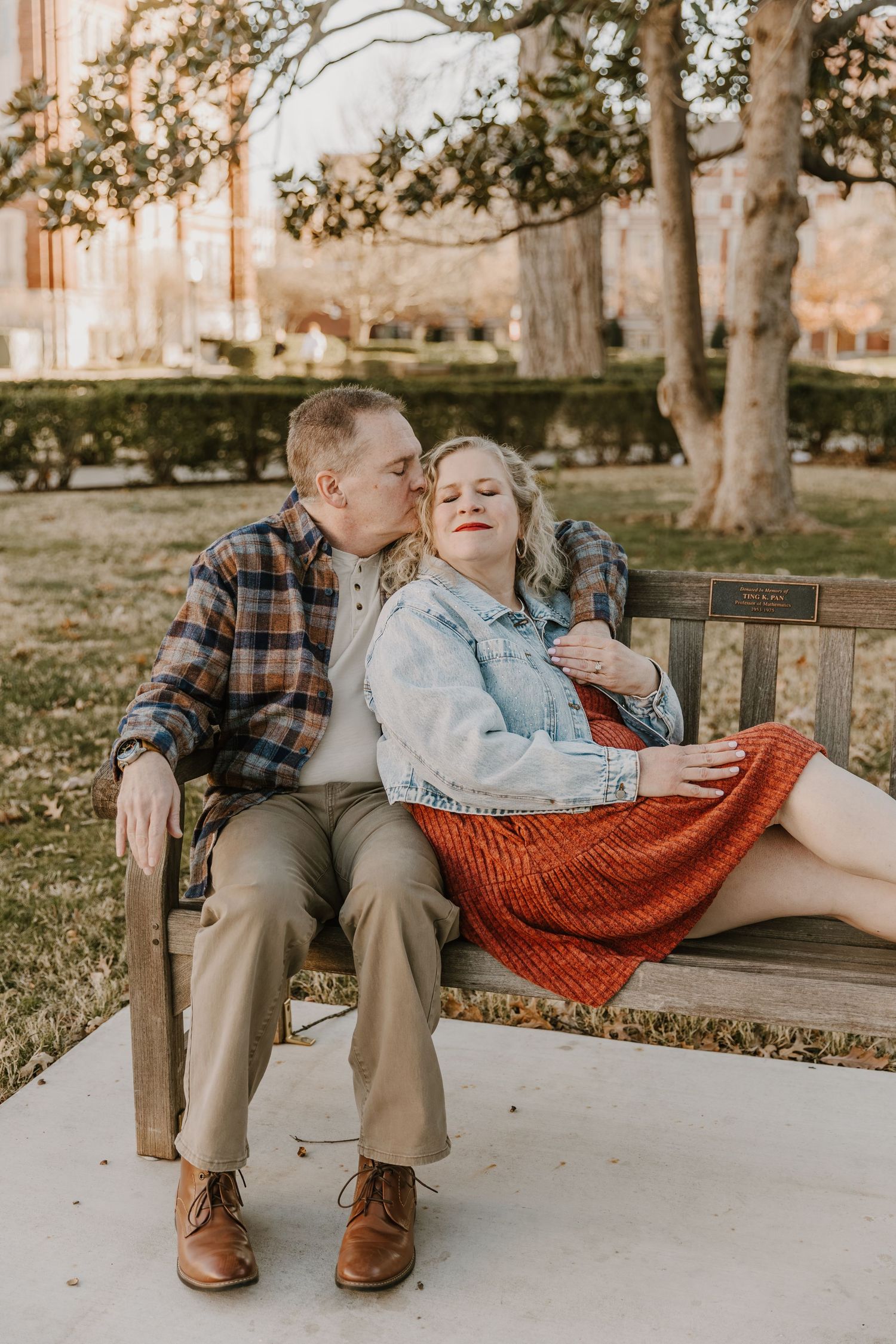 Couple sitting on a park bench, man kissing woman's cheek, she smiles in a red skirt and denim jacket.