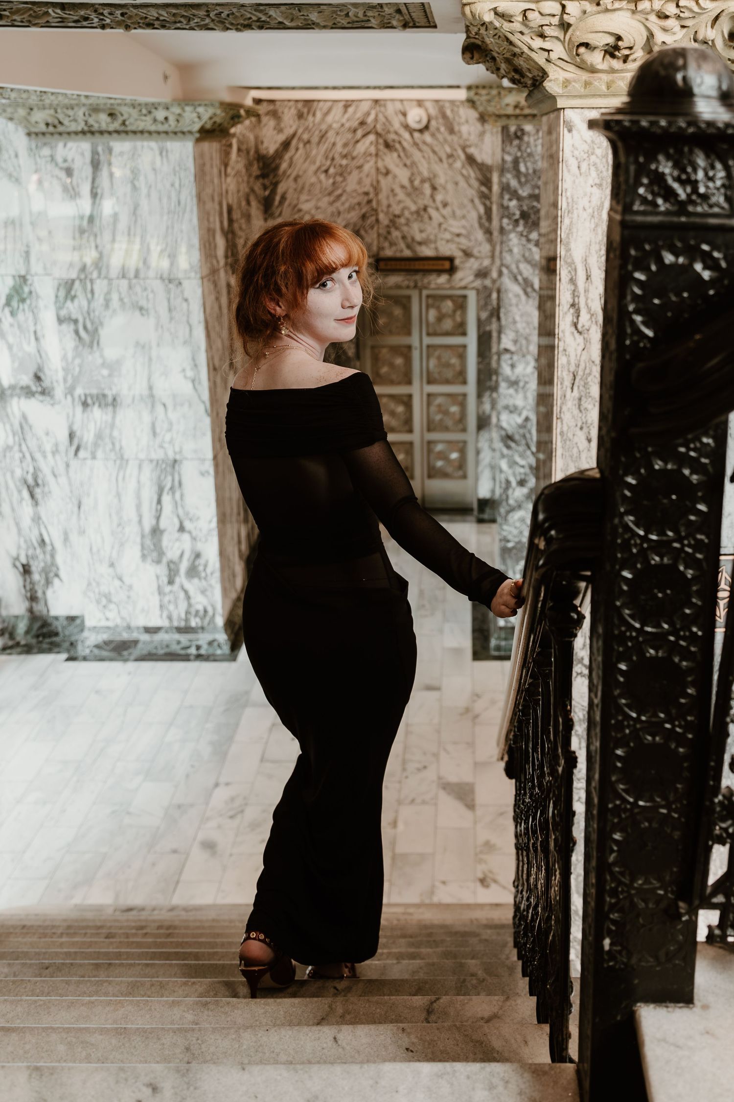 A person in a black dress poses gracefully in a vintage colonnade with white pillars and decorative wall details.