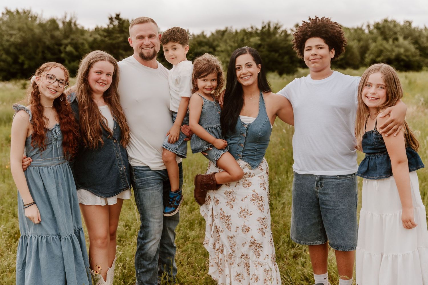 Happy blended family of seven posing together outdoors in a green field, wearing casual summer outfits.