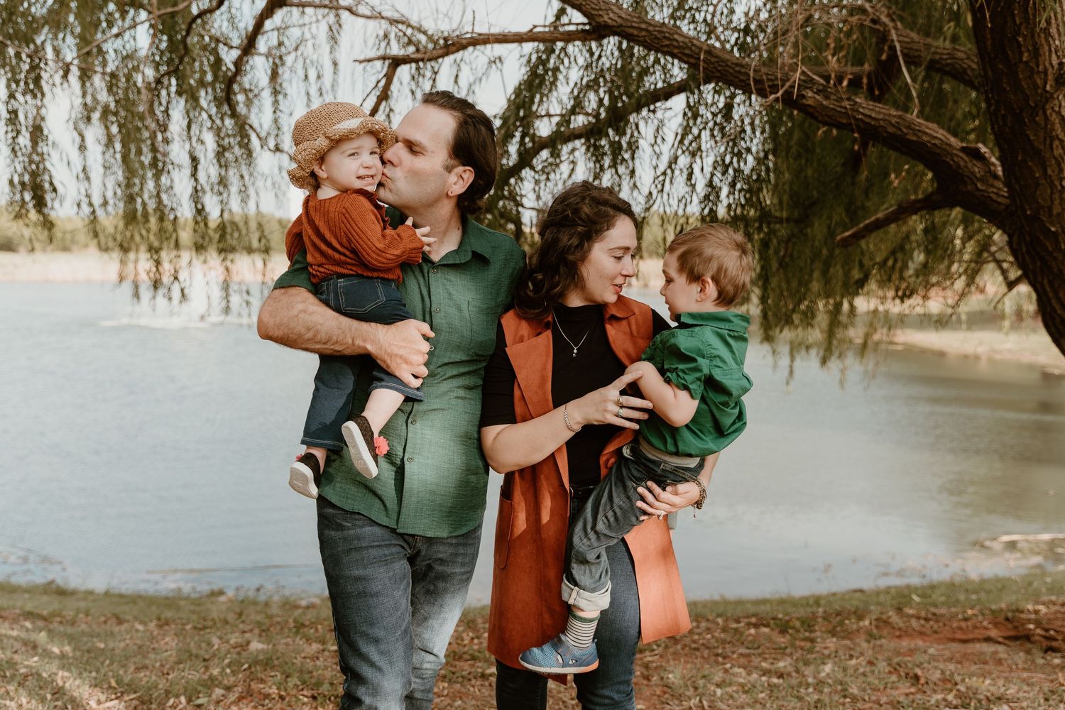 A family shares tender moments together in a natural outdoor setting with willow trees in the background.