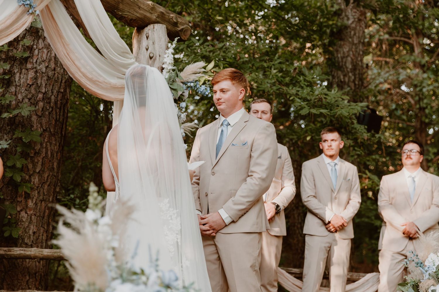 Wedding party wearing light beige suits and a flowing white wedding dress pose outdoors in a garden setting.