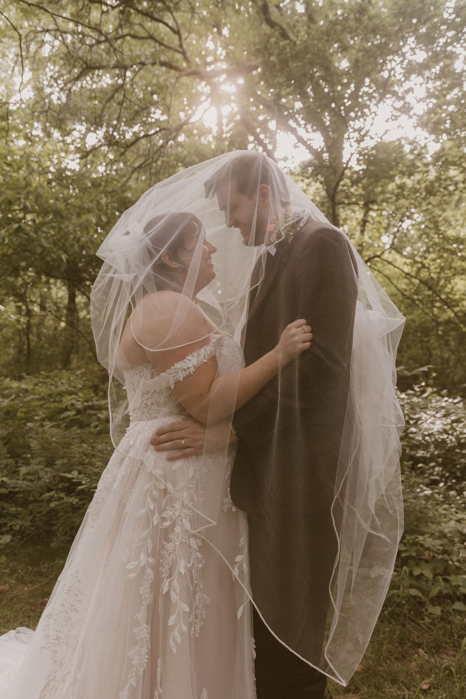 Bride and groom share romantic moment under flowing veil in sunlit forest, wearing lace wedding dress and brown suit.