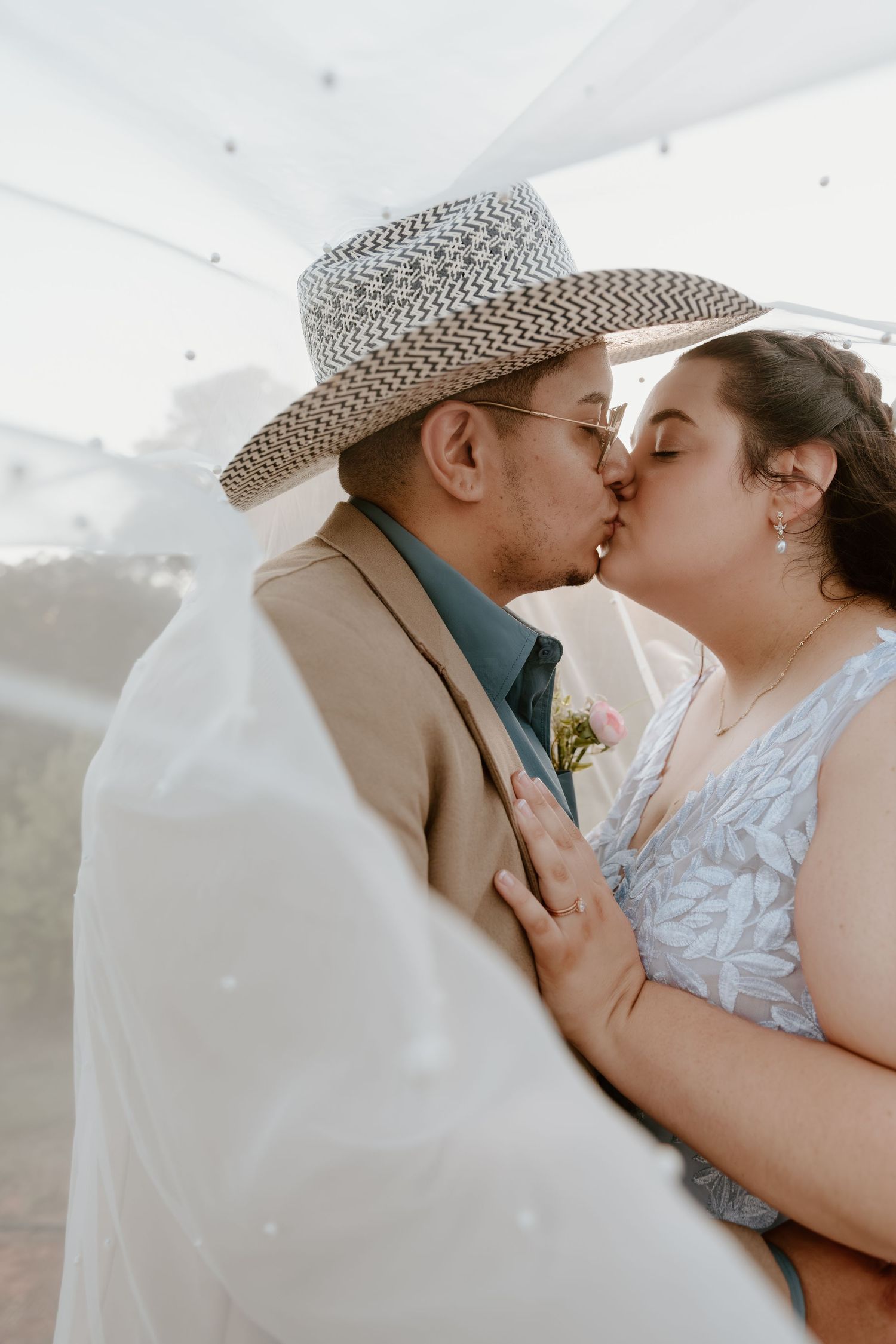 A couple shares a romantic kiss under a white tent at their outdoor wedding, the groom wearing a straw hat and tan suit.