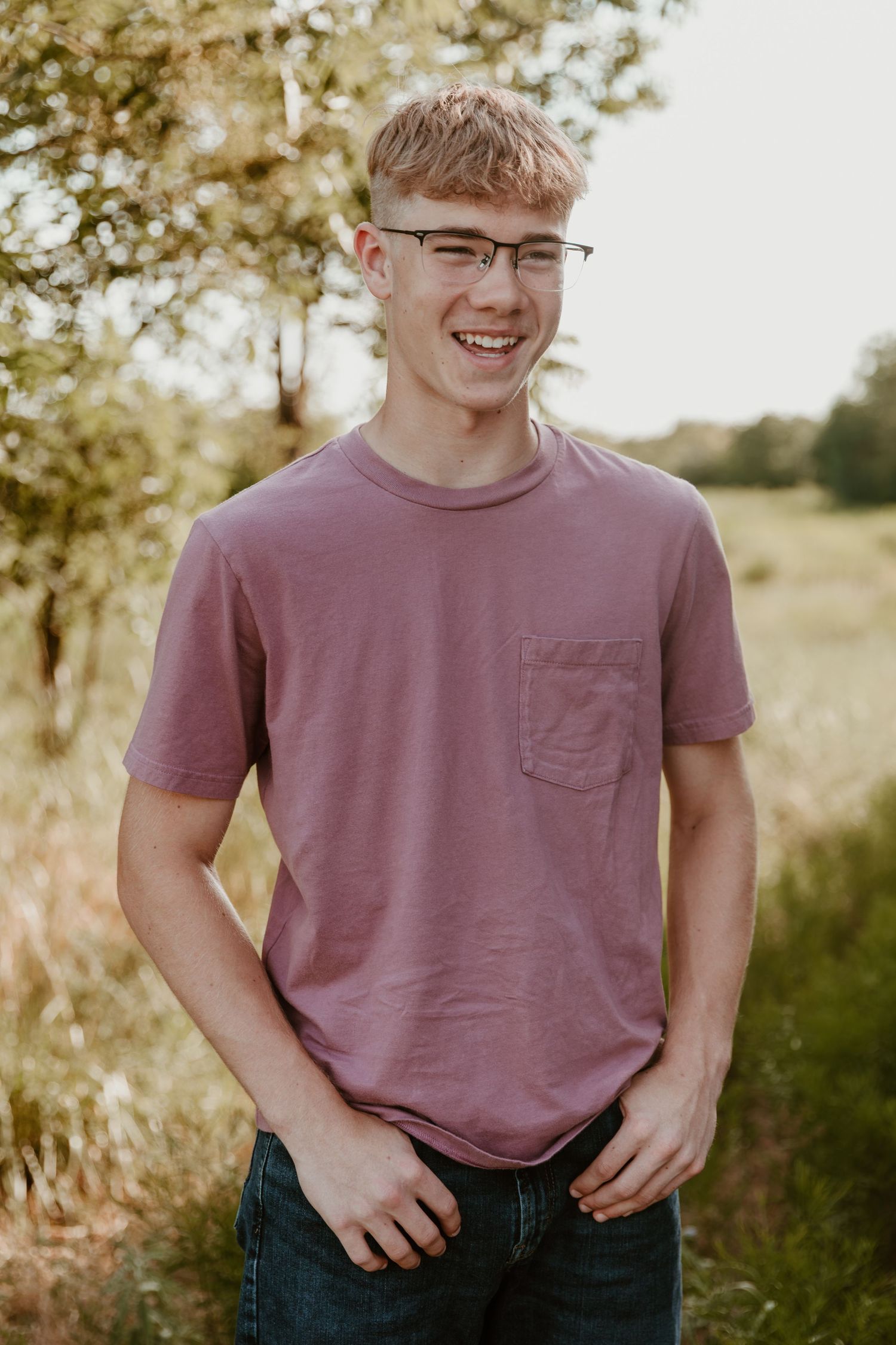 A person wearing glasses smiles outdoors in a mauve t-shirt and jeans against a soft natural background.