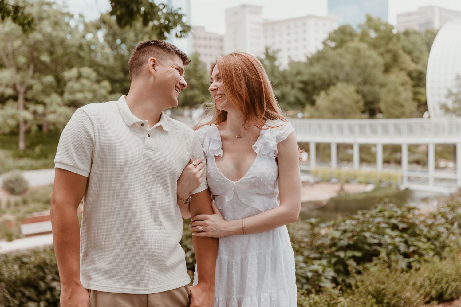 A romantic couple in casual summer attire share a laugh while standing in a lush park garden with urban buildings behind them.