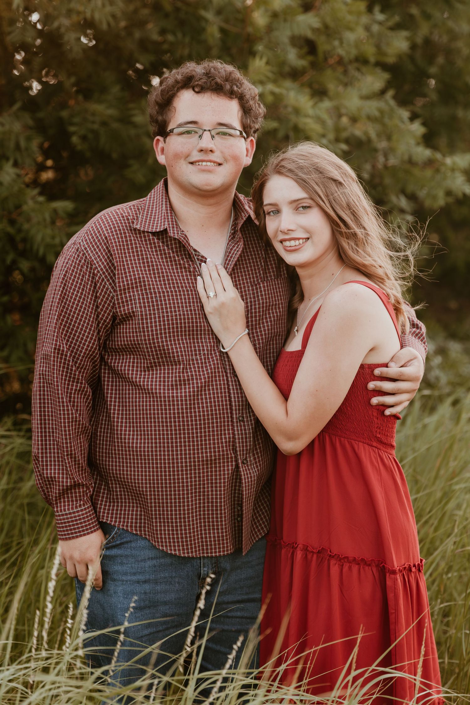 A romantic couple poses together in a field at sunset, with her wearing a red dress and him in a plaid shirt.