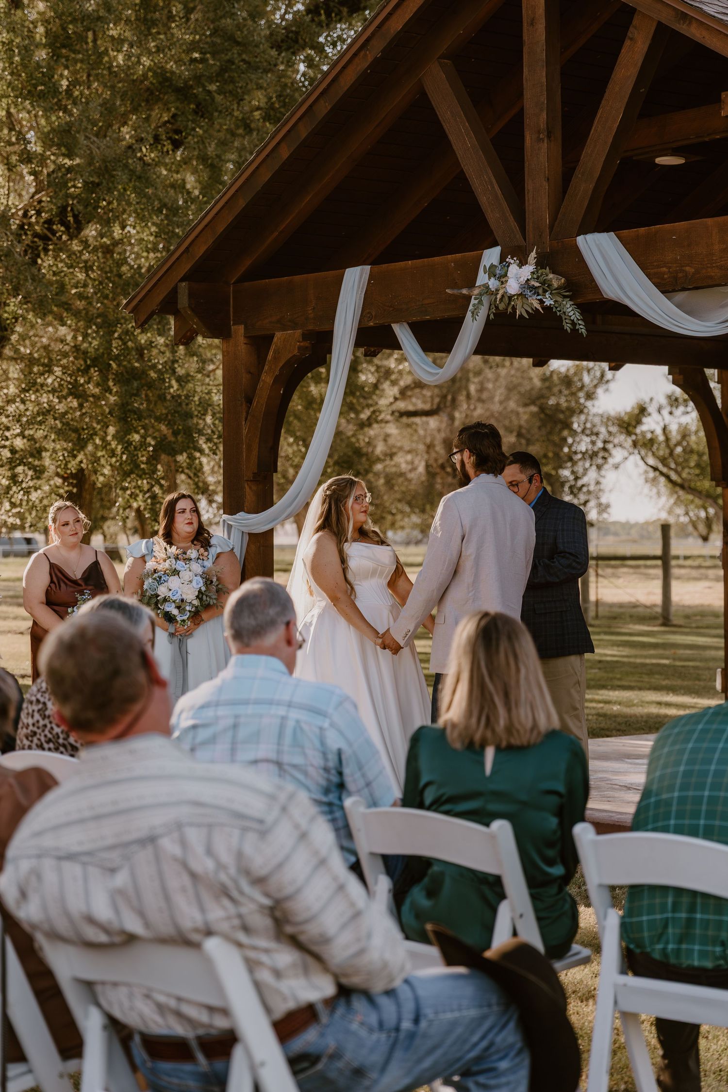 Wedding guests seated outdoors under a rustic wooden pavilion during a celebration ceremony.