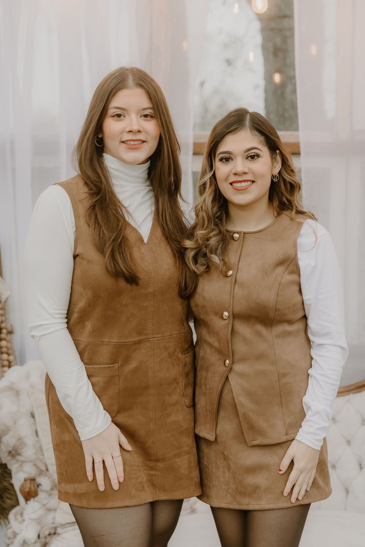 Two people wearing matching brown vests and white turtleneck sweaters pose together in a bright indoor setting.