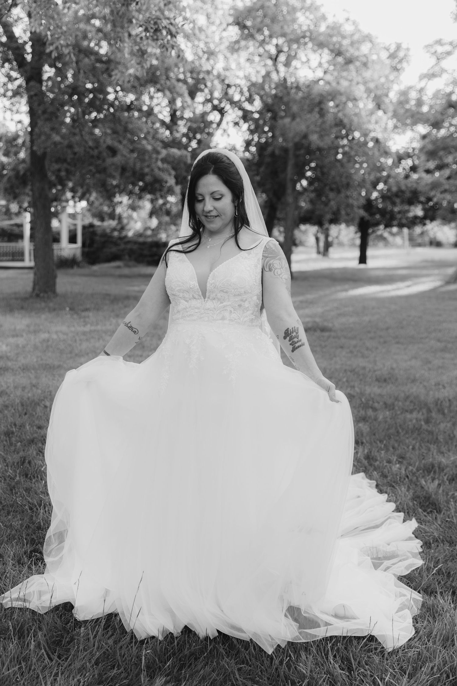 A bride in a flowing white dress sits gracefully on grassy ground during an outdoor photo session.