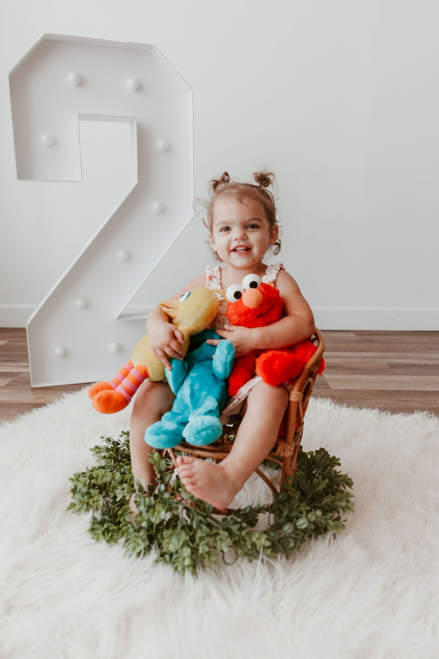 Happy toddler seated with plush toys next to large decorative number 2 on white fluffy rug with greenery wreath.