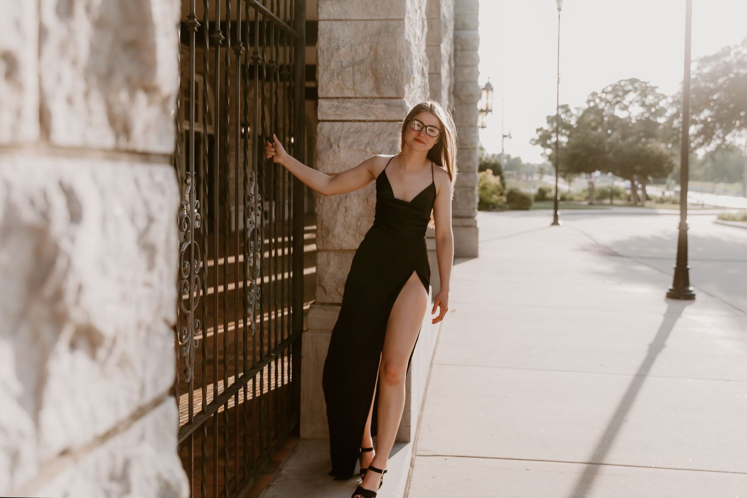 A person in a black dress with a slit leans against a stone wall with wrought iron gates during sunset.