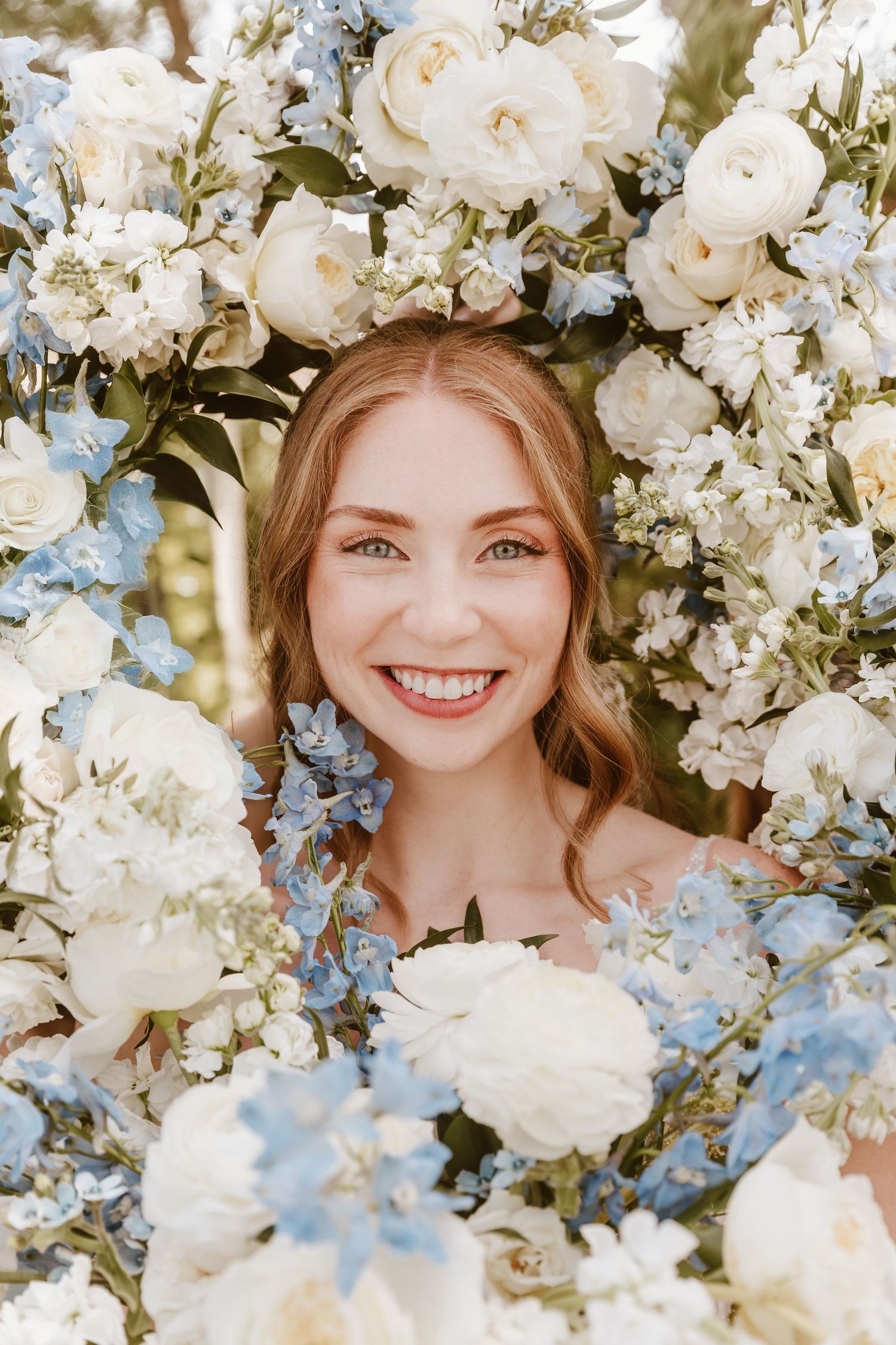 Smiling bride surrounded by lush white and blue floral arrangement including roses and hydrangeas.