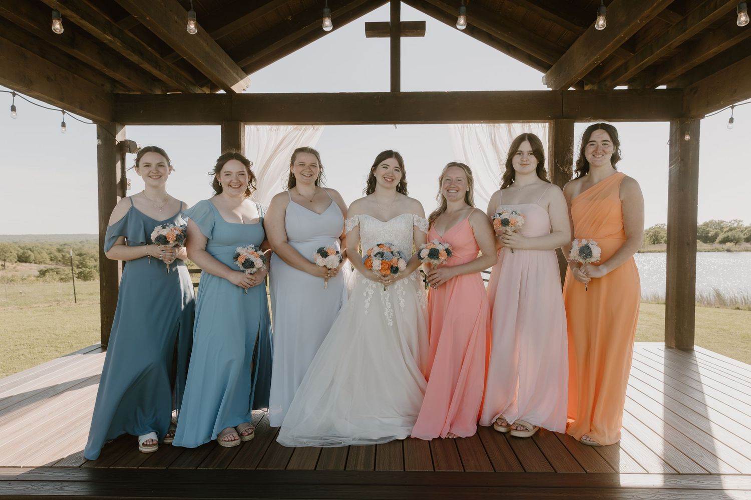 Bride and six bridesmaids in colorful mismatched gowns pose under a wooden gazebo at an outdoor wedding venue.