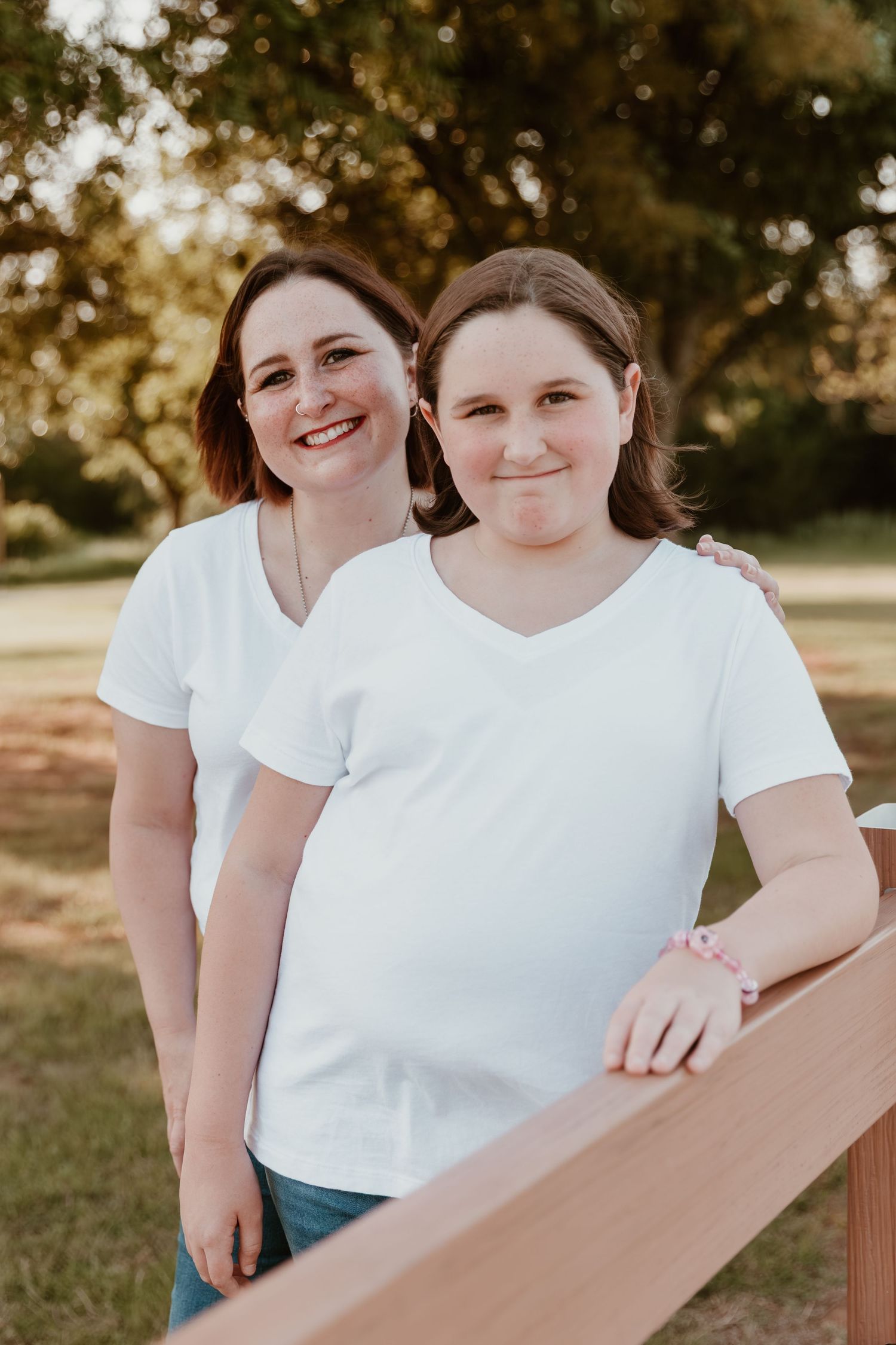 Two women in white t-shirts stand together outdoors at sunset leaning against a wooden railing.