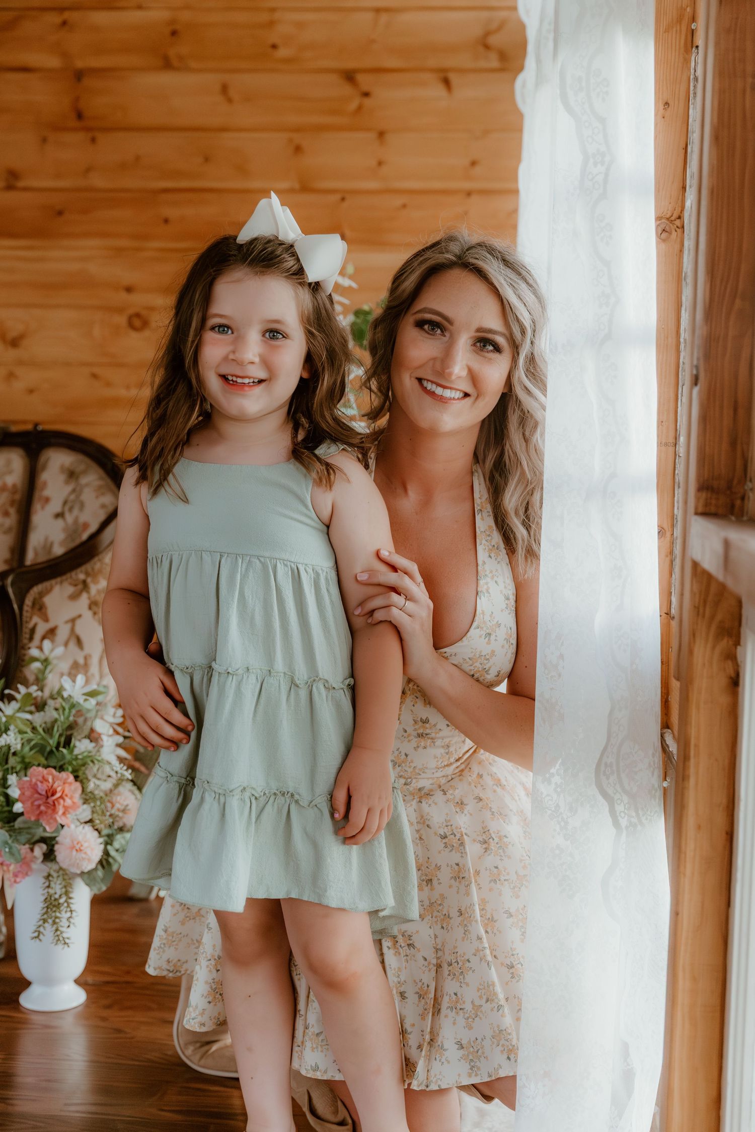 A tender moment captured in a rustic cabin interior with sage green and floral dresses beside a white curtained window.