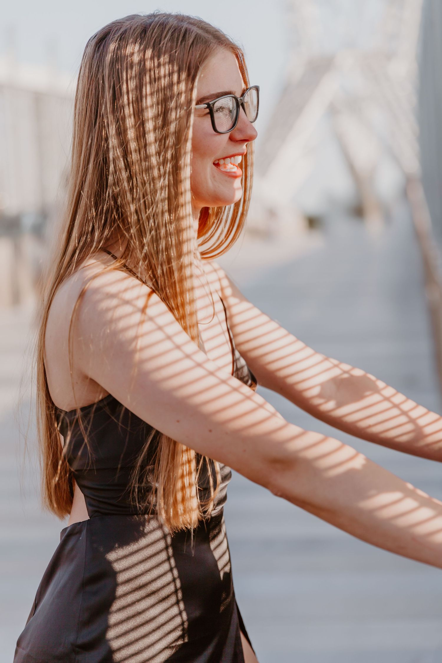 Sunlight creates striped shadows on a woman in glasses wearing stripes and dark clothing while reaching forward.