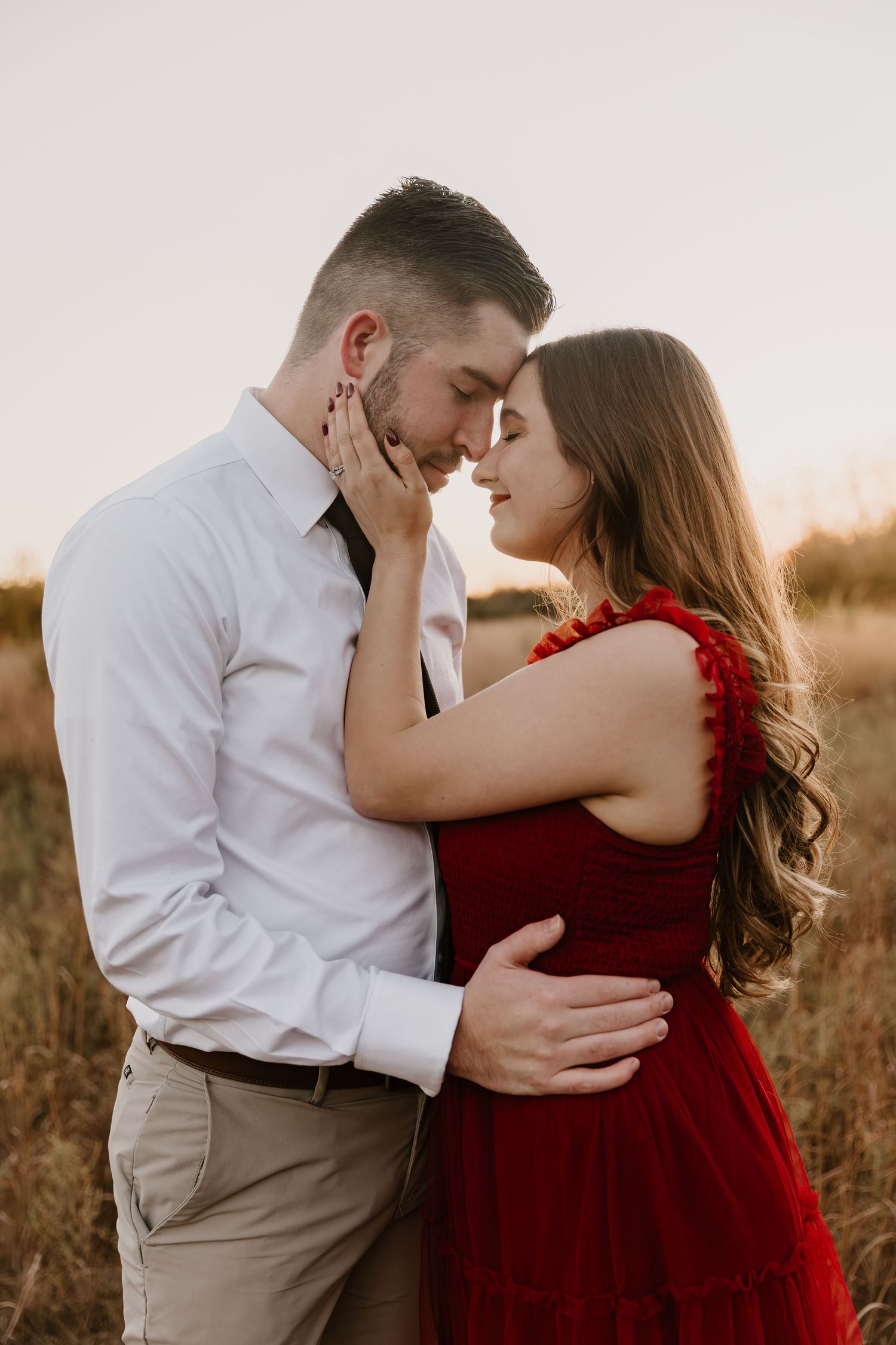 Romantic couple embraces in a field at sunset, with the woman wearing a flowing red dress and man in white shirt.