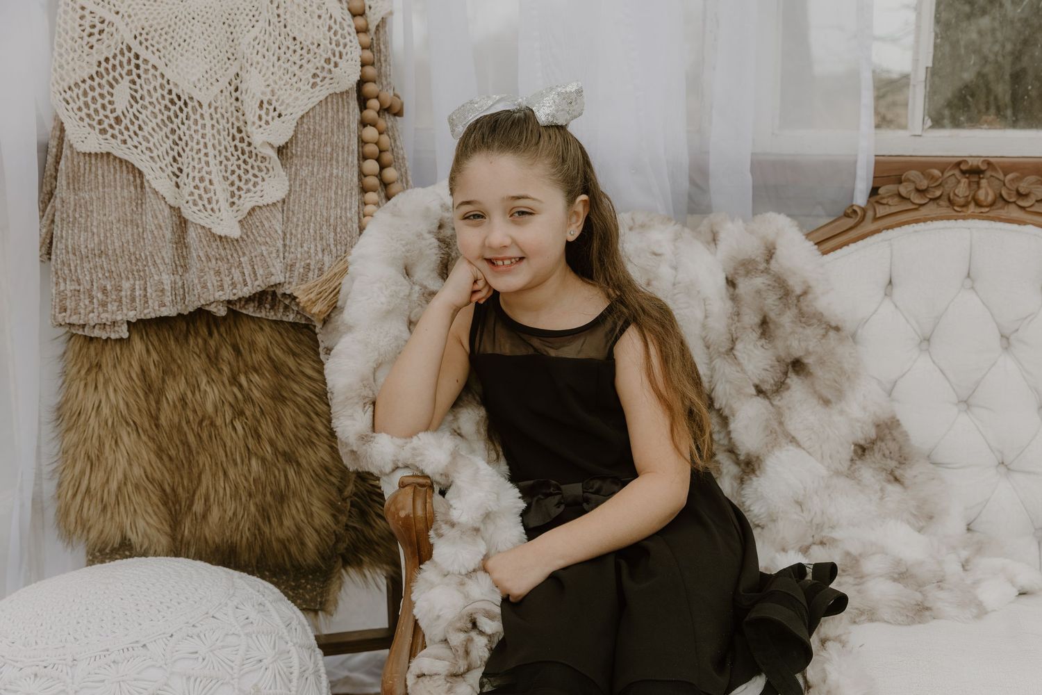 A young girl in a black dress sits elegantly on a plush white vintage sofa surrounded by decorative pillows and lace.