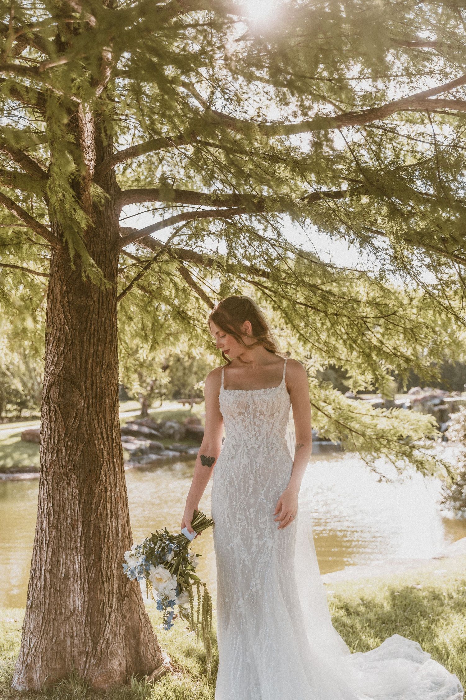 Bride in lace wedding gown holding bouquet stands under pine tree by serene pond in golden sunlight.