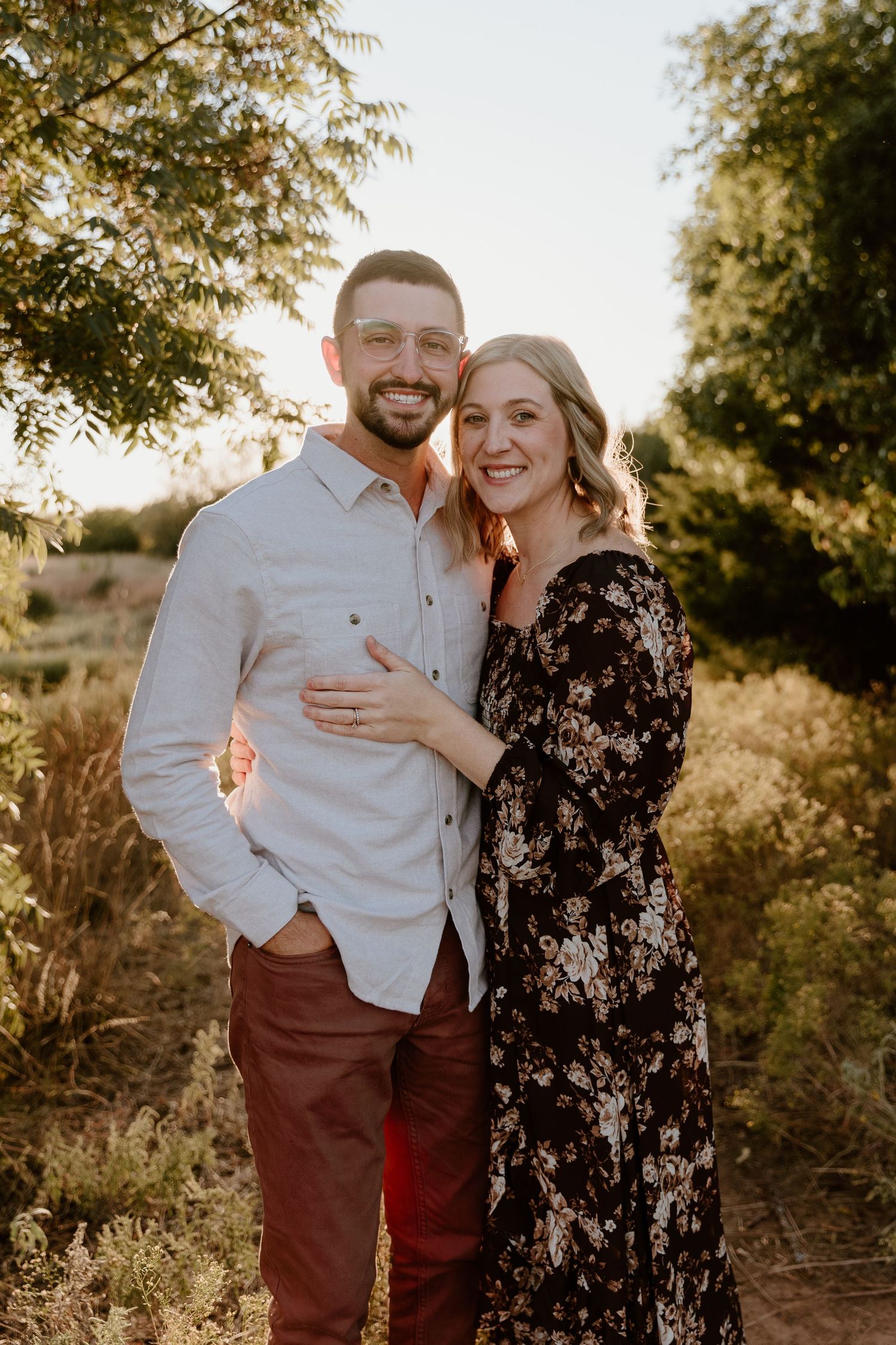 A couple embraces outdoors during golden hour with trees and sunlight in the background.