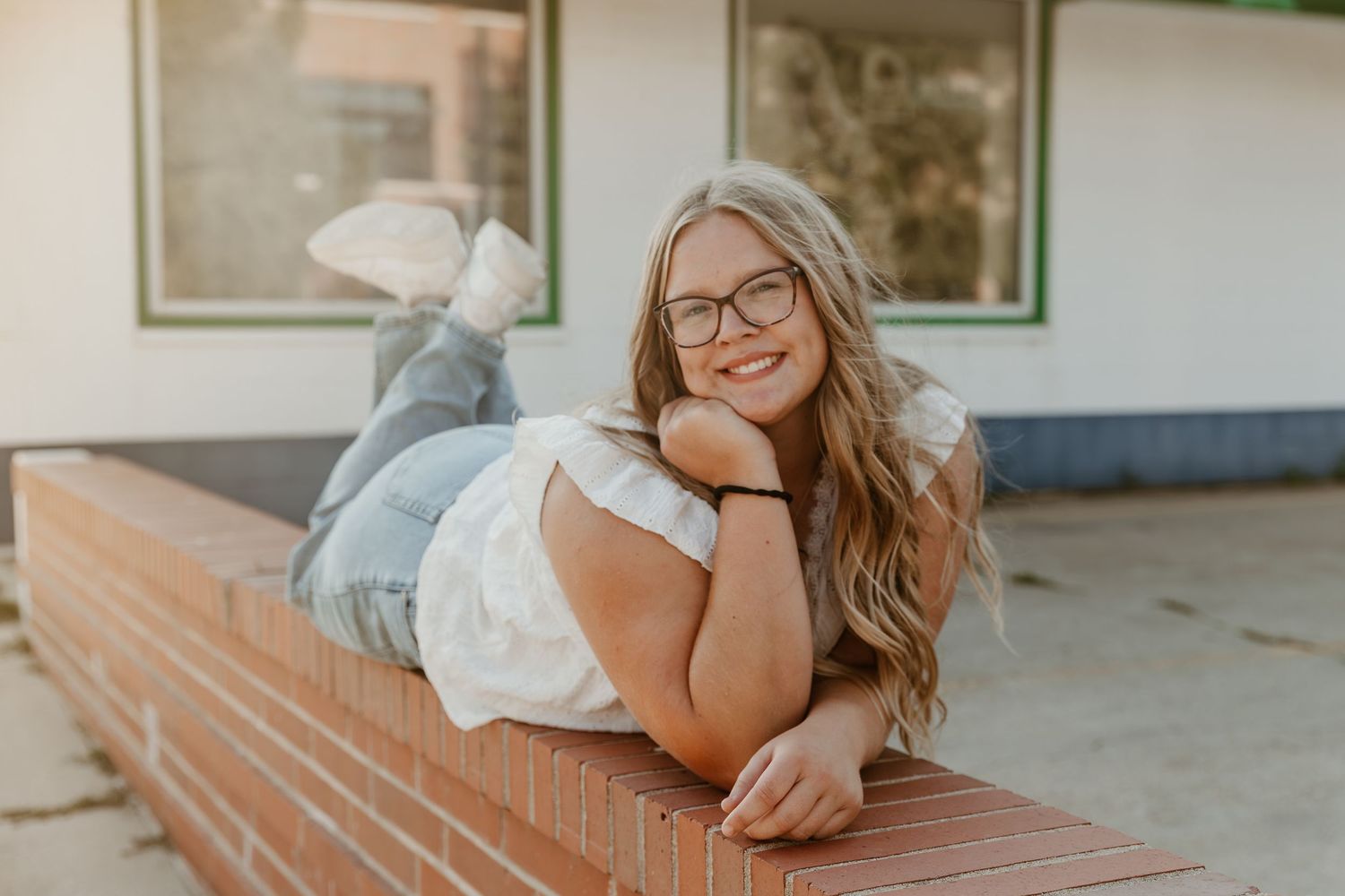 Young woman with glasses and long wavy hair lying casually on brick ledge while smiling at camera.