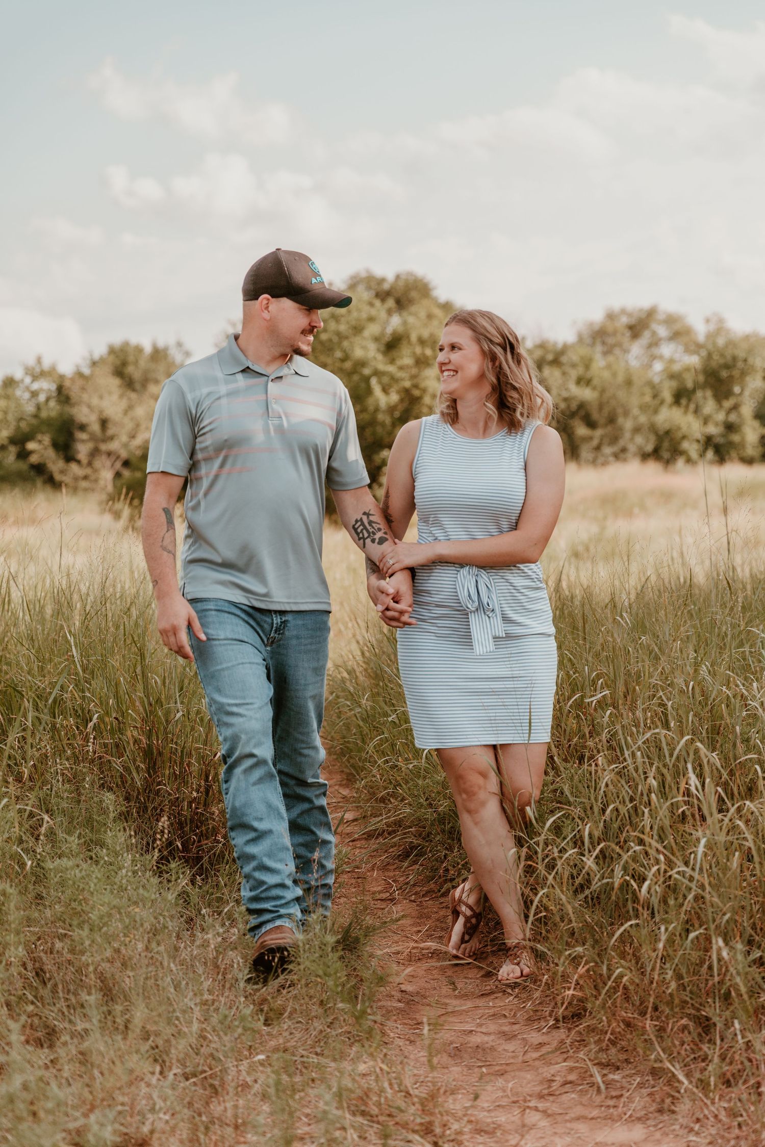 Couple walking together on a dirt path through tall grass in a golden-lit field at sunset.